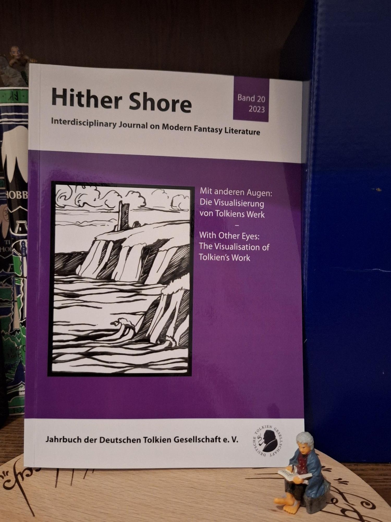 The cover of Hither Shore, Vol. 20: a purple cover with a black and white illustration of the hither shore. Next to it, the topic of the volume: "Mit anderen Augen: Die Visualisierung von Tolkiens Werk - With Other Eyes: The Visualisation of Tolkien's Work". The volume is framed by an Ü-Ei Bilbo and Gollum.