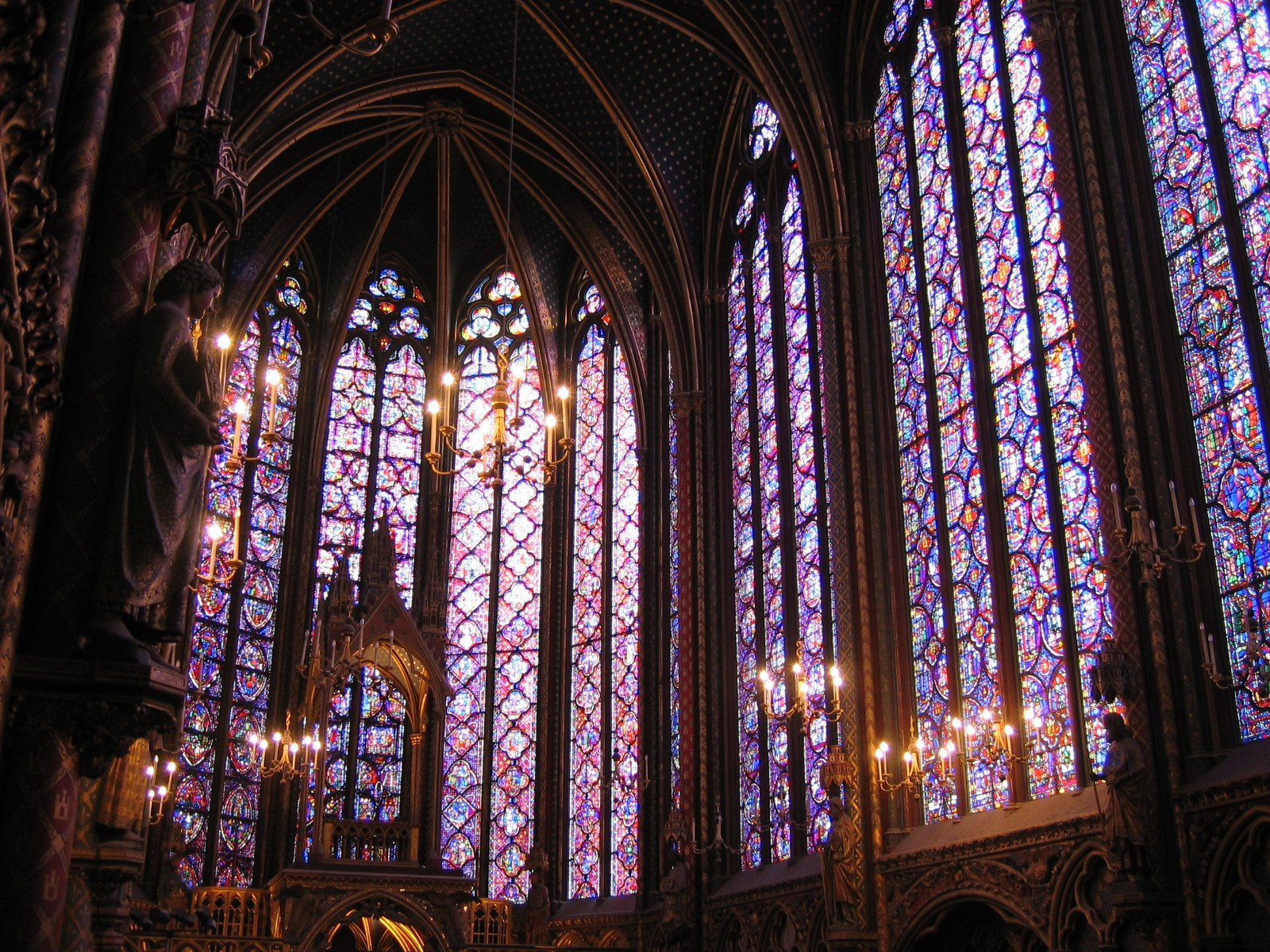 An interior photo of the stained glass in the Sainte-Chapelle chapel in Paris.  There are candelabra visible in front of the beautiful glass that towers overhead.  Purple and blue dominate the glass.