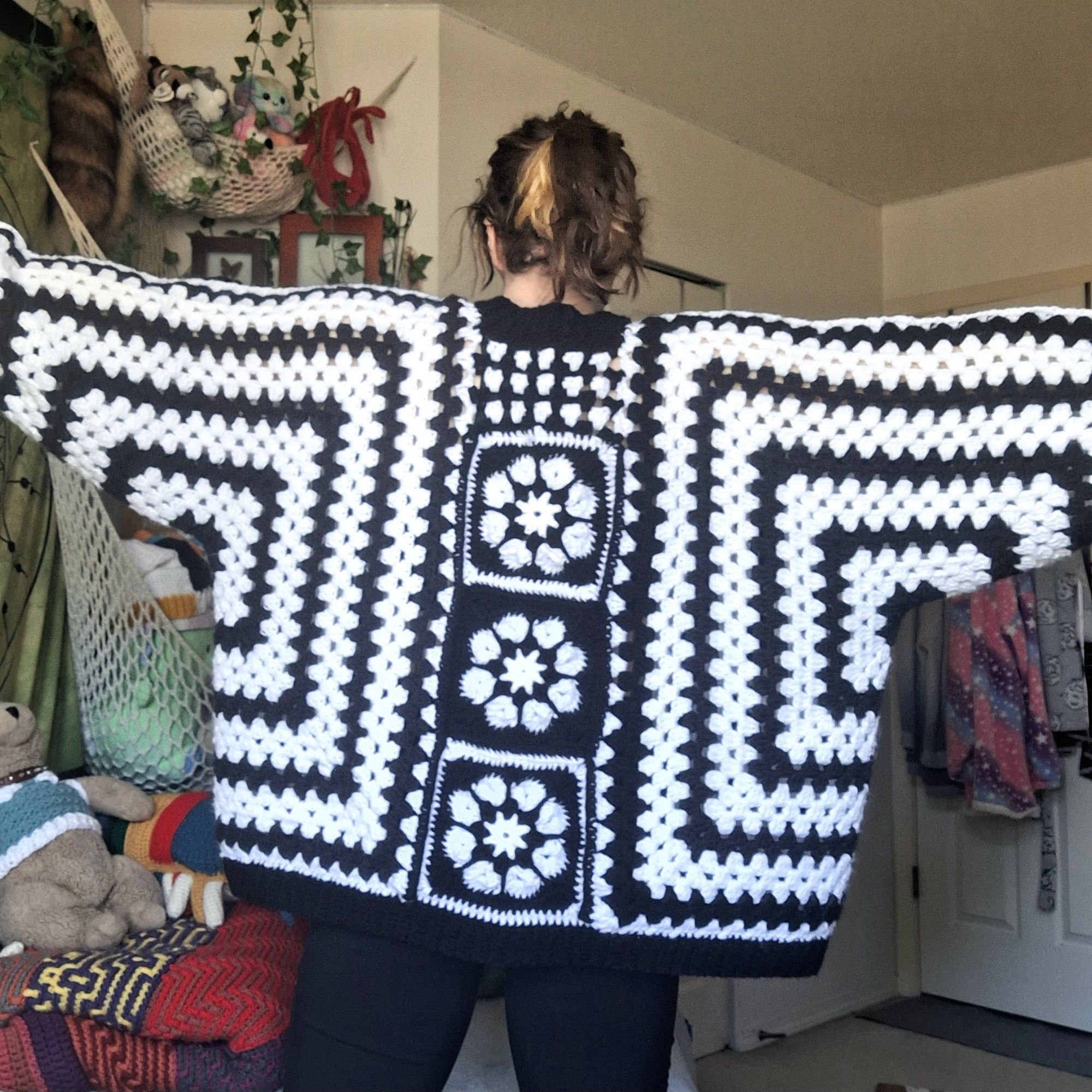 Photo of a person wearing a crochet sweater, showing the back of it. it has alternating black and white horizontal and vertical stripes, with 3 black and white flower granny squares down the middle.