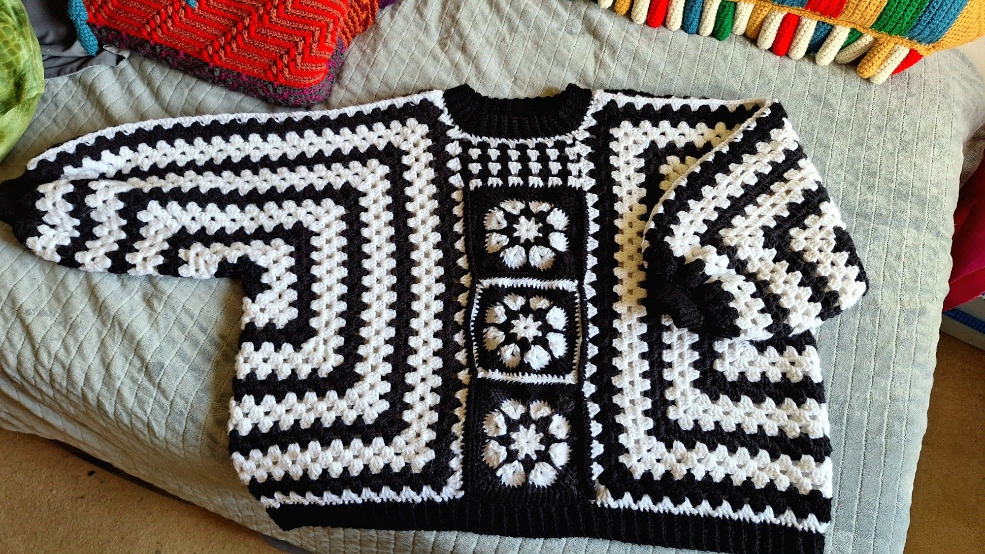 pictures of a black and white crochet sweater laying on a bed with one arm slightly folded. it has alternating black and white horizontal and vertical stripes, with 3 black and white flower granny squares down the middle.