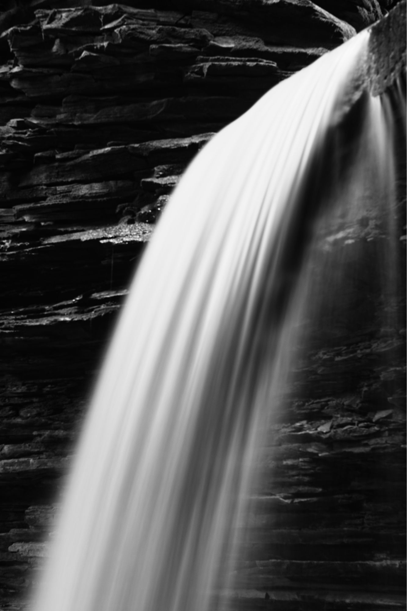 A black and white long exposure photograph of a narrow waterfall showing the slimy smoothness of the water against the dark jagged rock behind.