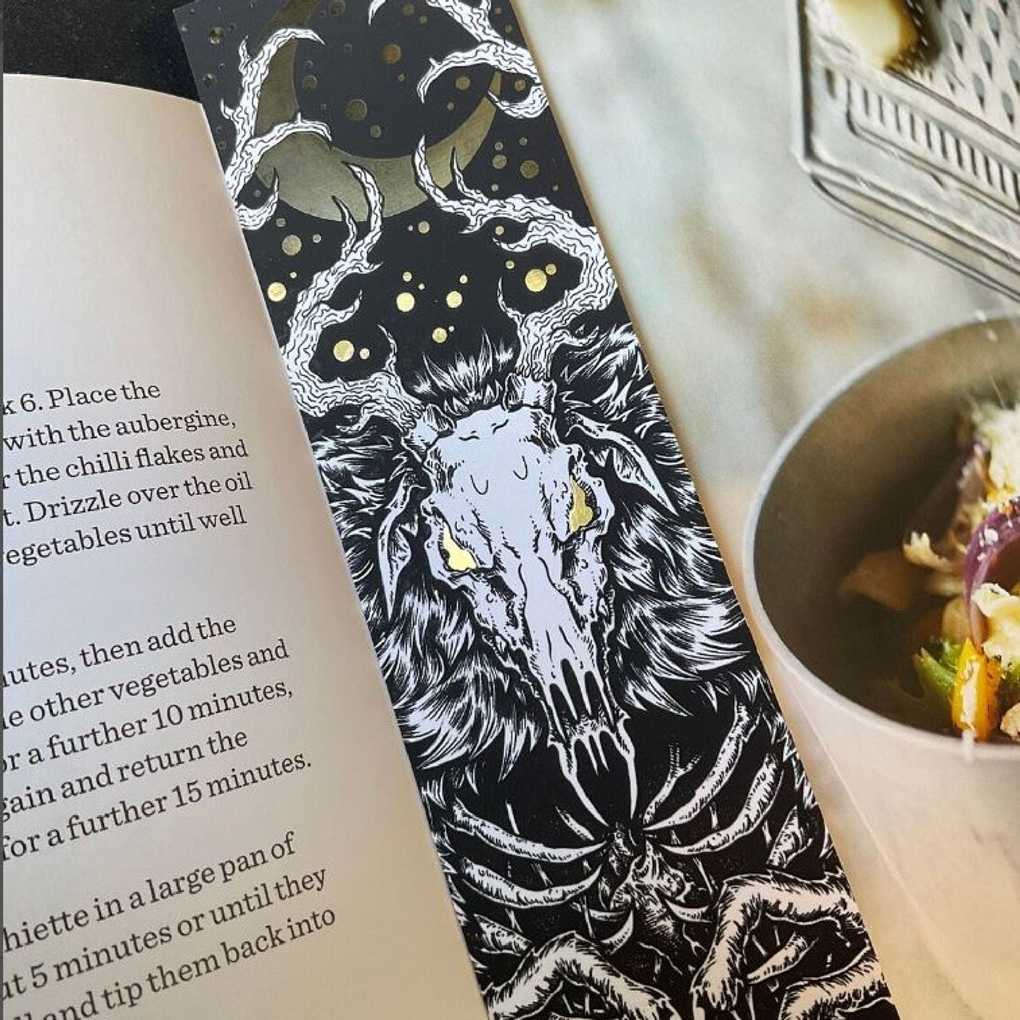 A photo of a large bookmark featuring a Wendigo. The design includes gold foil detailing, and is symmetrical.