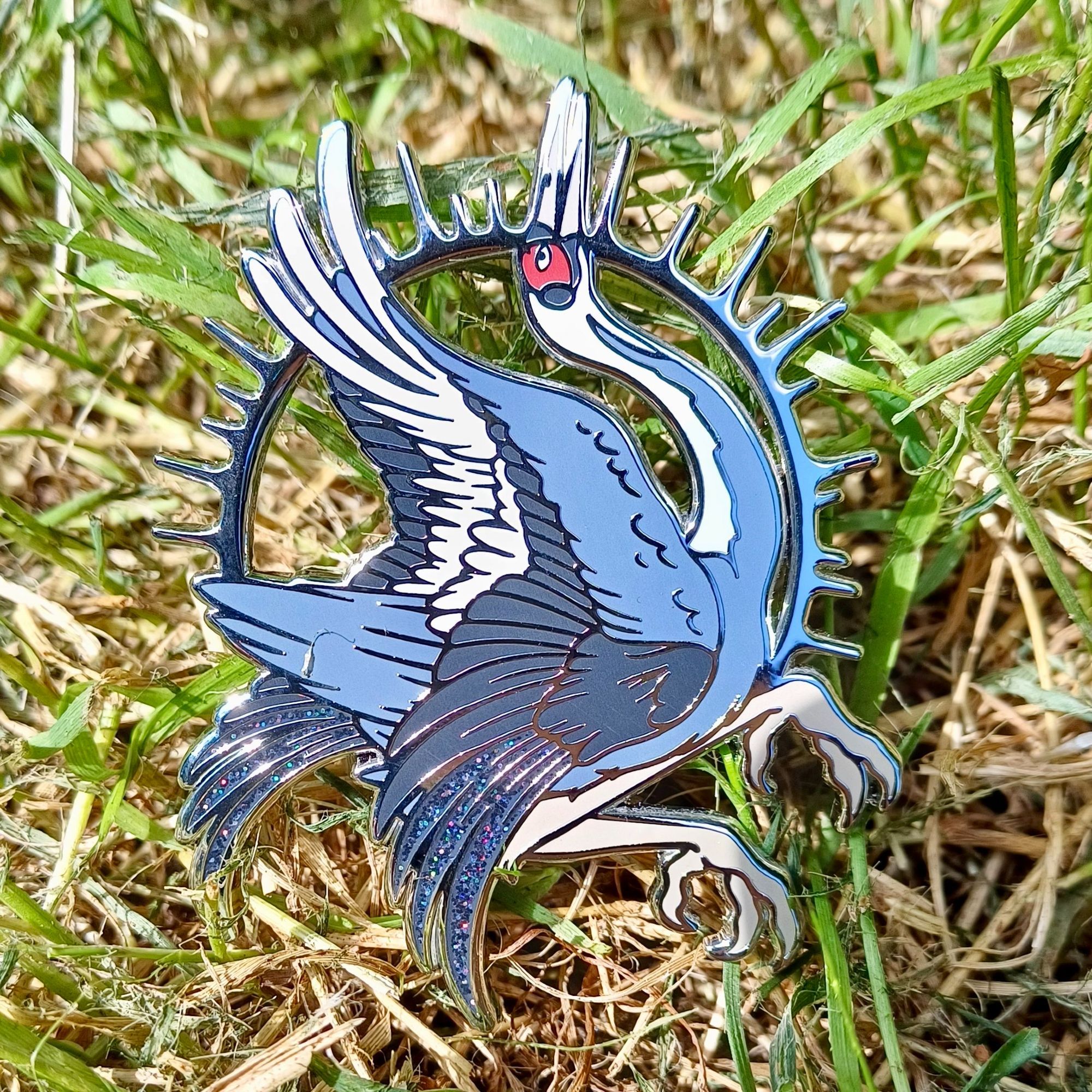 A photo of a large enamel pin badge featuring a White Naped Crane. The crane is in an arched and dynamic pose with it's wings out, and there's black glitter enamel detailing on the wing tips.