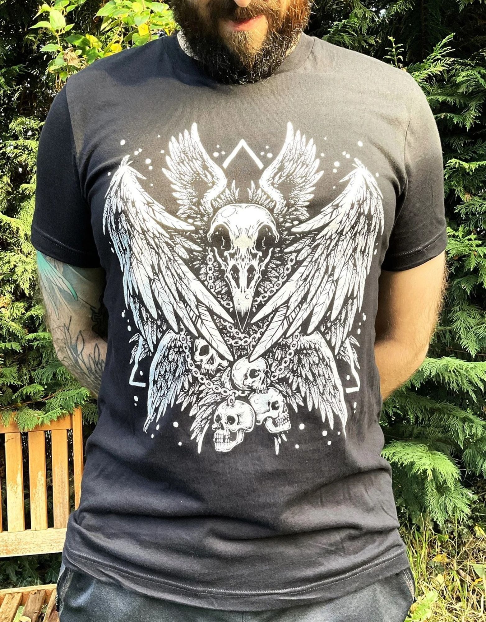 A photo of a model wearing a black t-shirt which features a seraphim bird skull design. The design is symmetrical and entirely screenprinted in white, and features feathered wings surrounding a large bird skull. There are some smaller human skulls chained around it's neck too.