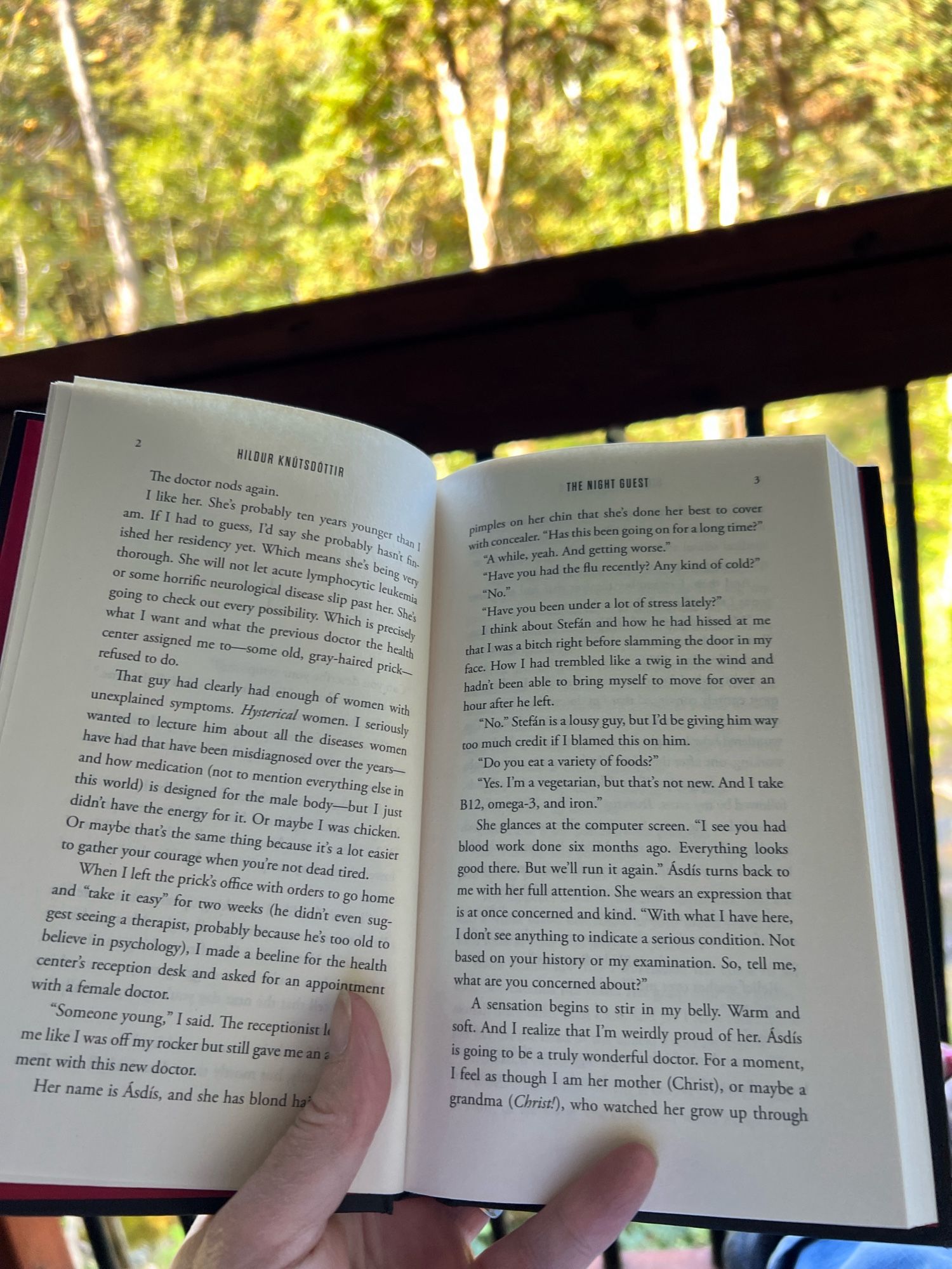 A hand holding open a book, with deck railing and green trees in the background