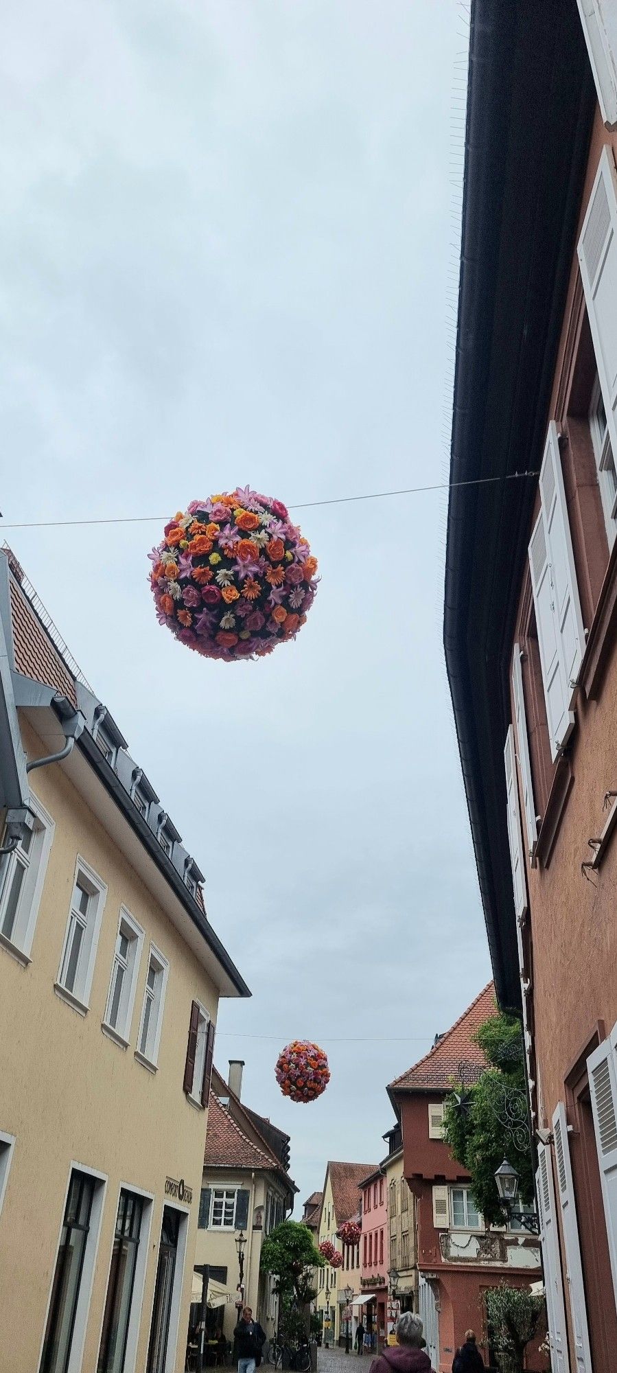 Bunte Blumenkugeln an Drahtseilen hängen über einer Straße mit älteren Häusern.