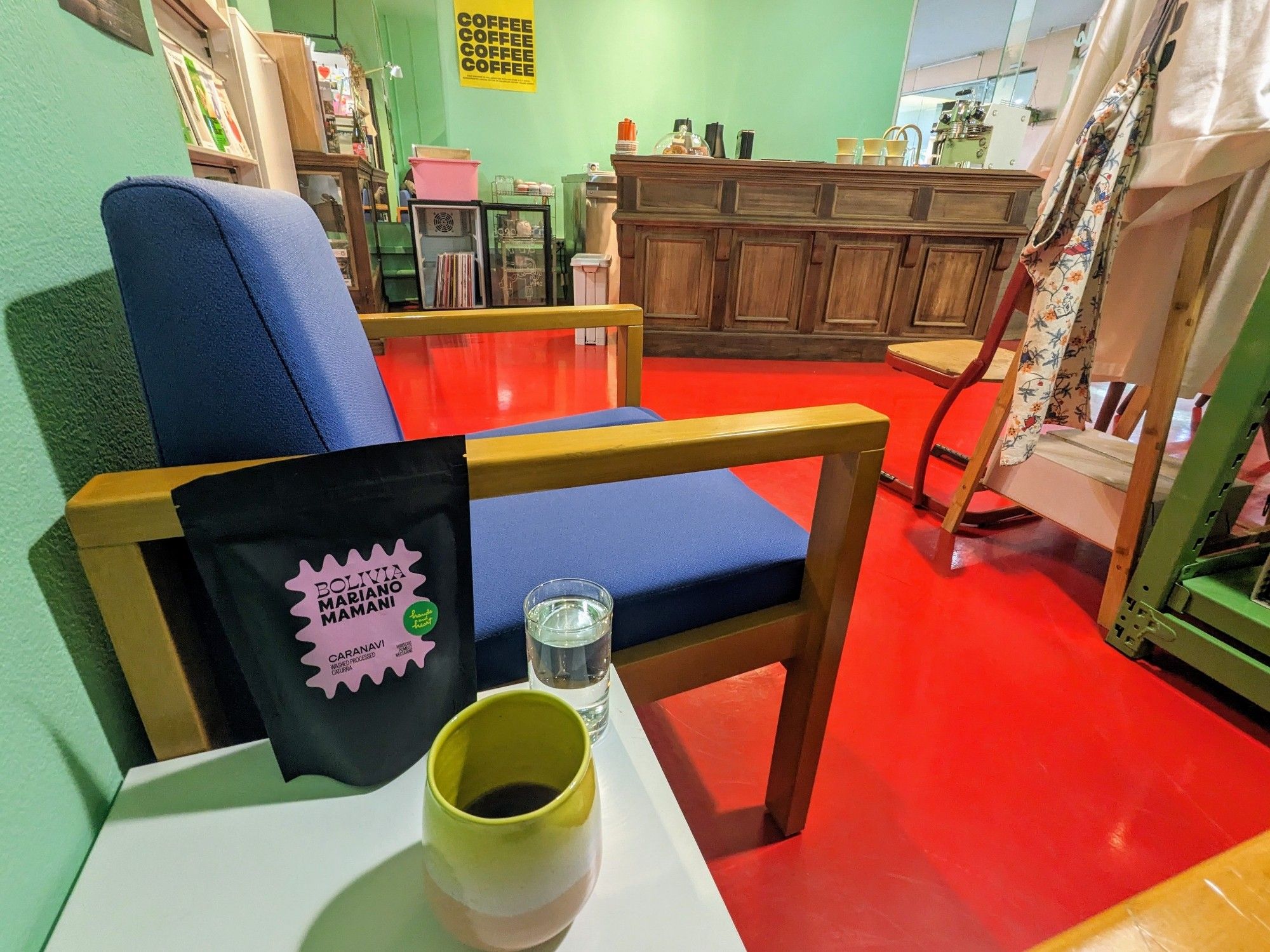 A wide-angle shot inside Hands and Heart Coffee Roasters in Bangkok, Thailand. Visible on the table in front is the aforementioned pourover coffee in a colorful glazed-pottery cup, with a bag of beans and a glass of water behind. The remainder of the shot shows the bright, arty, colorful shop interior and the paneled-wood brew bar.