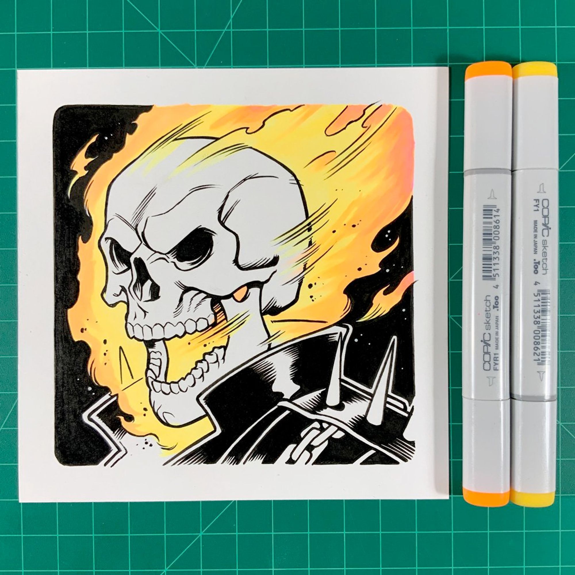 A drawing of Marvel Comics' Ghost Rider on a square piece of Bristol paper with two Copic markers sitting to the right.  The drawing is in black and white except for the flames on Ghost Rider's head which are in bright yellow and orange.