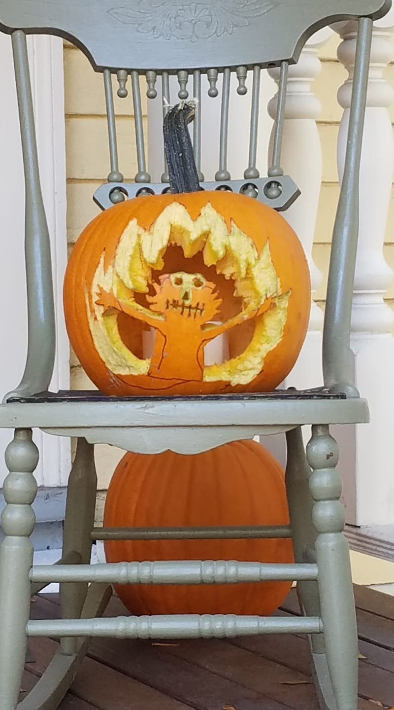 A carved pumpkin sits on a rocking chair. It is carved with the visage of the flaming Elmo gif. Another, uncarved, pumpkin sits on the porch