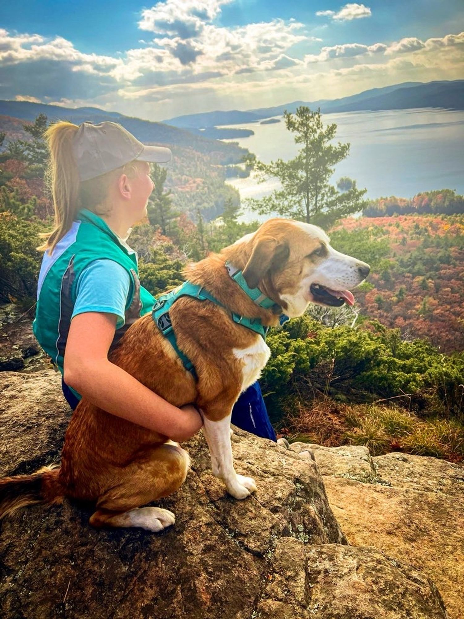 A woman and her dog sit on the edge of a cliff, looking away from the camera at the scene in front of them. The woman has her right arm over the dogs torso in a sign of affection. The woman is looking forward while the dog is looking slightly to the right. In front of them is a large lake dotted with small islands. Mountains can be seen on the opposite shore, and the sky above is blue with many clouds. Directly below them is a little bit more of the shoreline; some of the trees still retain their colorful fall leaves