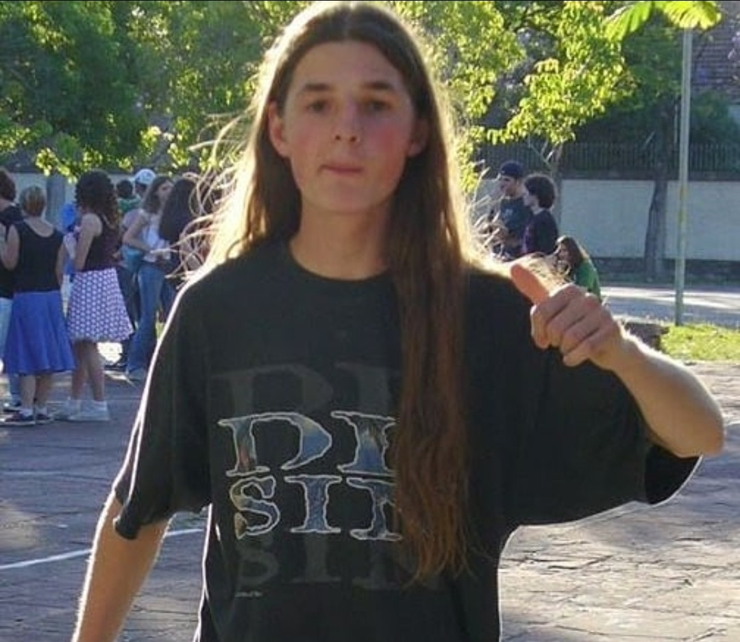 Foto do Fane jovem. Camiseta preta da banda dr. Sin, cabelo cumprido com cobrindo parcialmente o grande logo da camiseta. Fazendo joinha com o mão e no fundo dá para ver uma movimentação d pessoas em um recreio de colégio. 