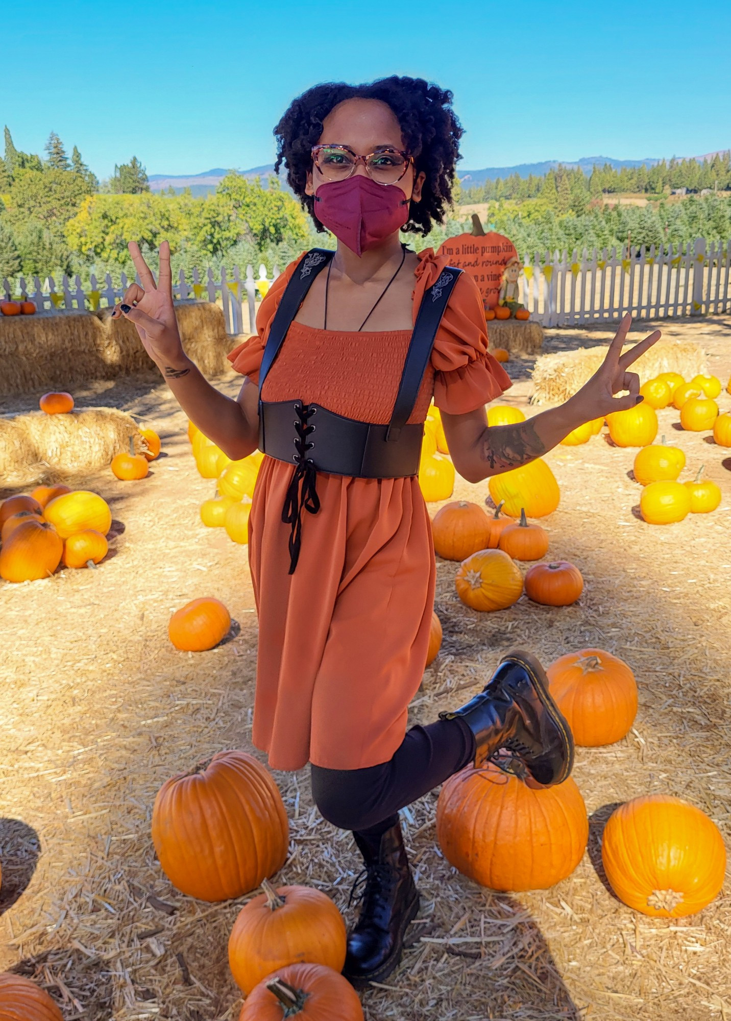 AmethystMoon101 is wearing a burnt orange dress, a black harness, black leggings, black boots, a burgundy mask and orange retro cat eye glasses. She's surrounded by lots of orange pumpkins and haystacks. She's posed with both her arms out and both hands throwing up peace signs. One leg is lifted and kicked out a little. 