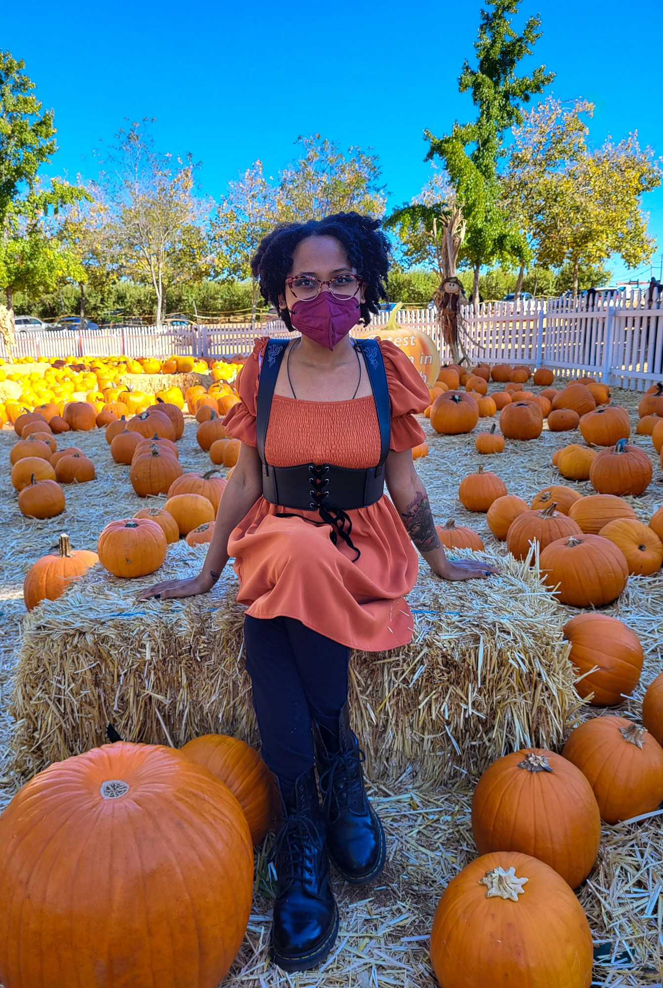 AmethystMoon101 is sitting on a hay stack, surrounded by a bunch of orange pumpkins. She's wearing a burnt orange dress, a black harness, black leg
gings, black boots, a burgundy mask and orange retro cat eye glasses. She's staring directly at the camera while both hands are laying on the hay stack. One of her legs is crossed over the other one. 