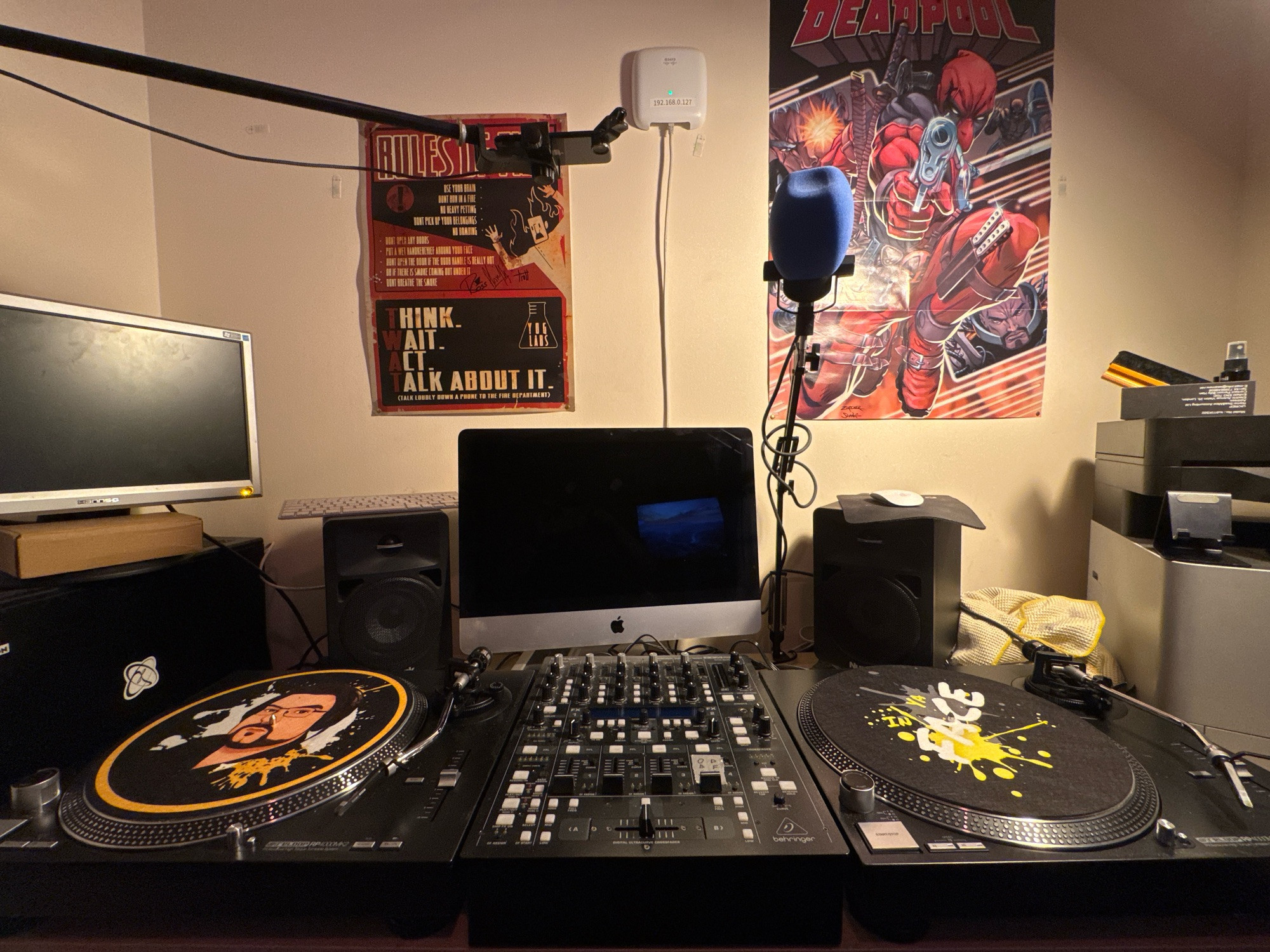 two Reloop turntables, a Behringer DDM-4000, an iMac, a pair of Numark monitor speakers, and a PodMic on a swing arm