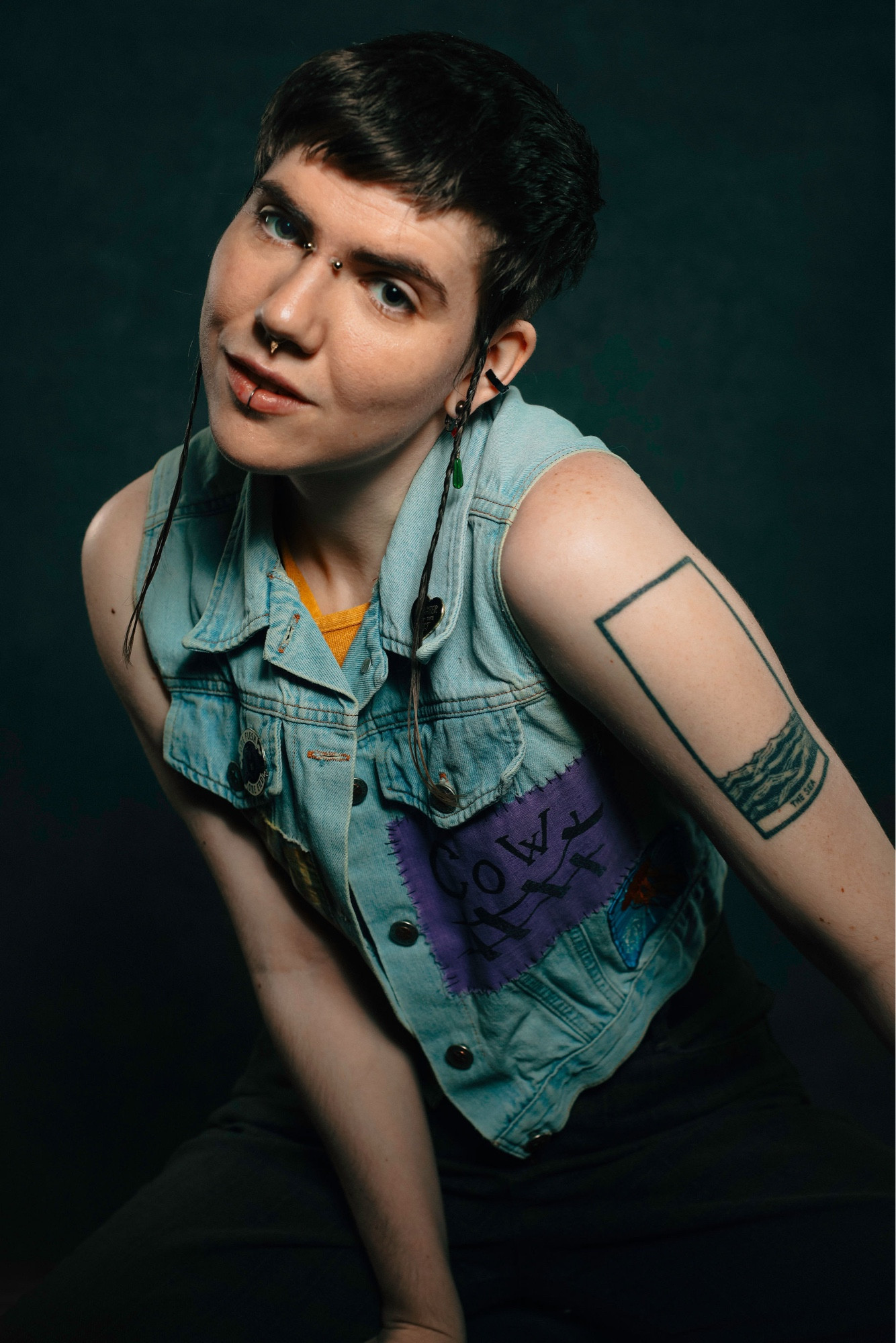 A photo of Sea, a white trans person leaning sideways and smiling at the camera. They have short borne hair, a myriad of piercings and tattoos, and wear a jean vest.