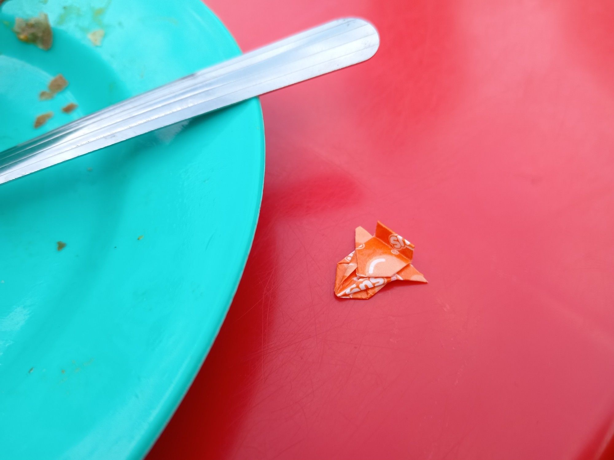 Red plastic tabletop, teal plate on left third of photo, a tiny orange origami aircraft on the table.