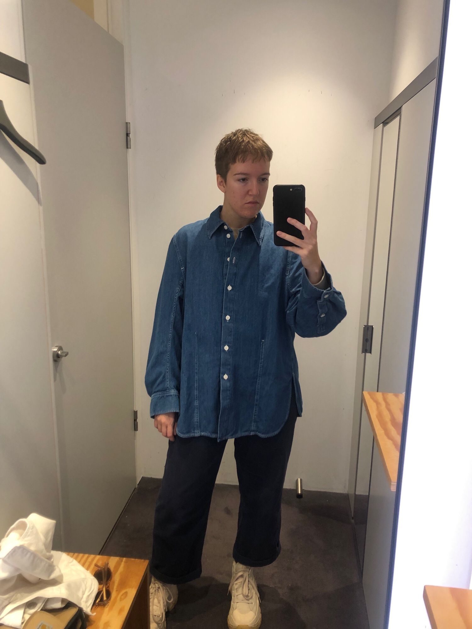 A mirror selfie of Hollie in a changing room mirror. They are wearing blue trousers, white trainers and a crisp new long denim shirt. The shirt is buttoned almost to the top, it has structured lines and a curved hem. 
