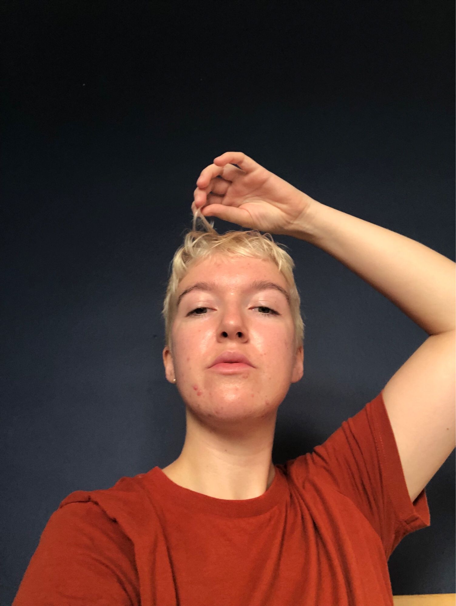 Hollie in a red T-shirt against a dark navy wall. They have their arm above their head, playing with freshly dyed bleach blonde hair