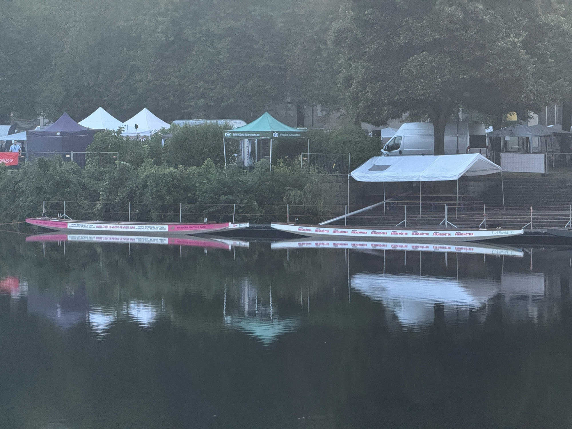 Drachenboote liegen im Wasser und dahinter stehen aufgestellte Faltpavillons