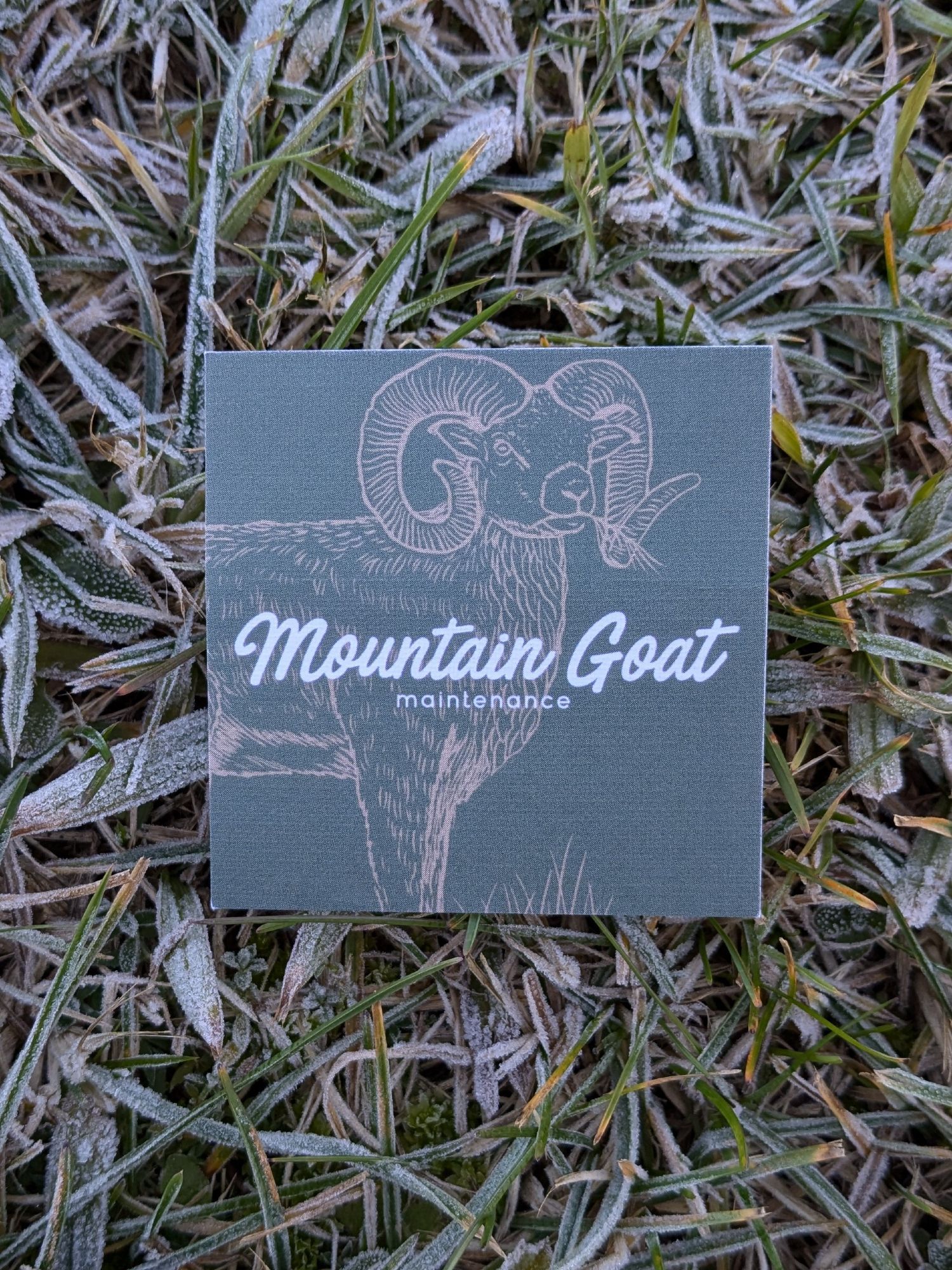 A green business card resting on frosty grass. The card reads Mountain Goat maintenance - white text over a yellow outline of a goat.