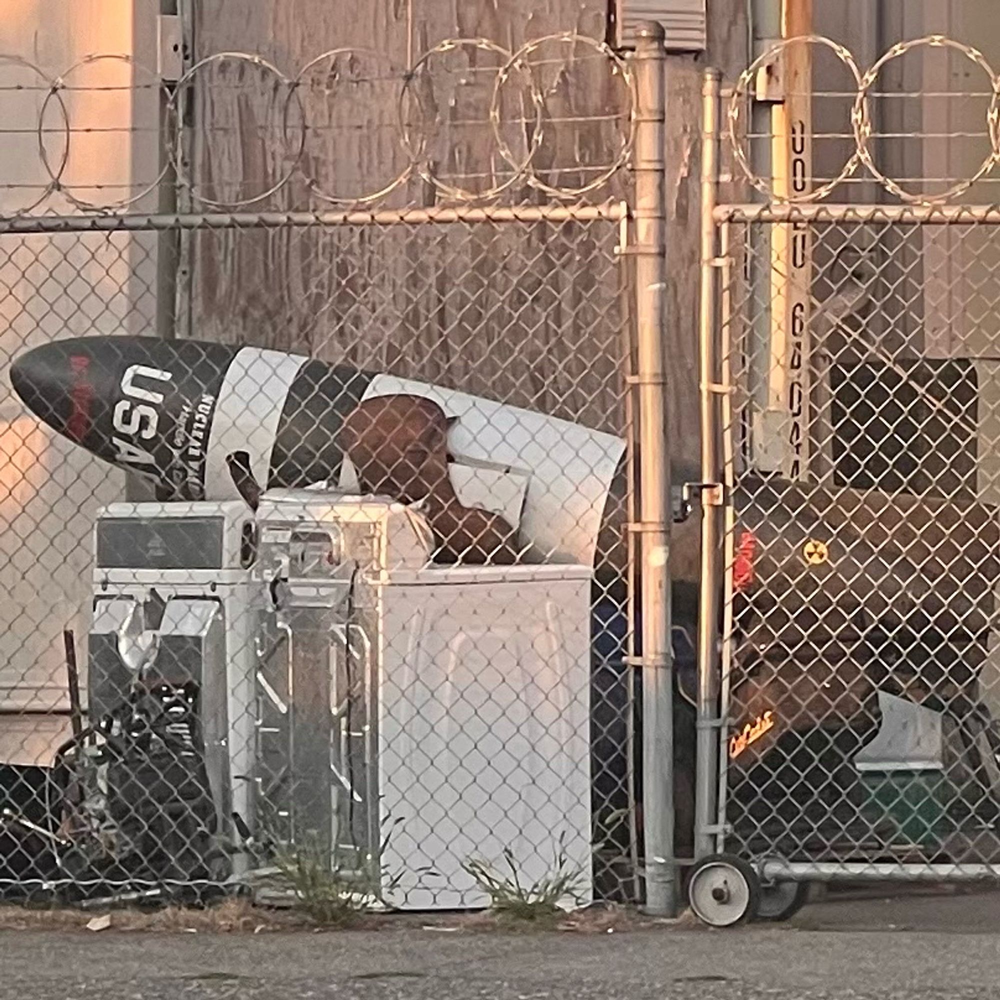 A large very bomb shaped item leaning on top of a derelict washer and dryer outdoors beside a chain link fence with an extremely threatening looking barbed wire top