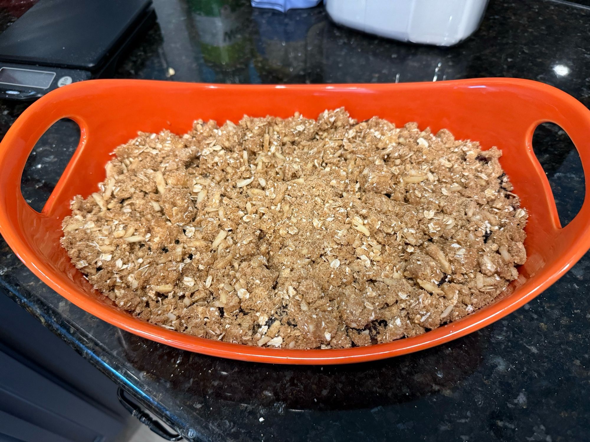 An orange colored oval baking dish filled with the fruit completely covered with light brown oat and almond crumble topping 