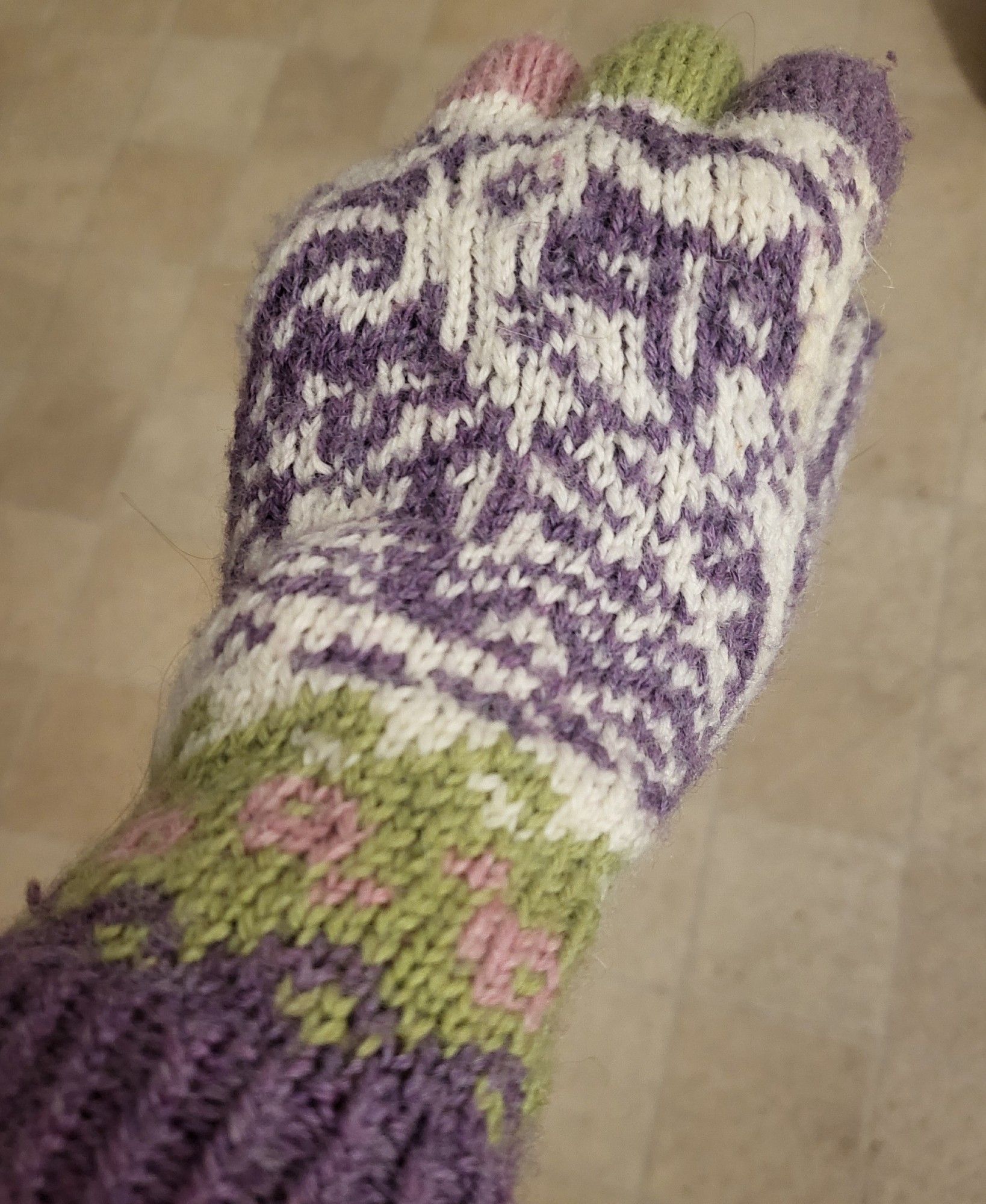 A hand knitted glove with purple, green and pink fingers and cuff, and in the hand part a cat outline in purple on a white background.