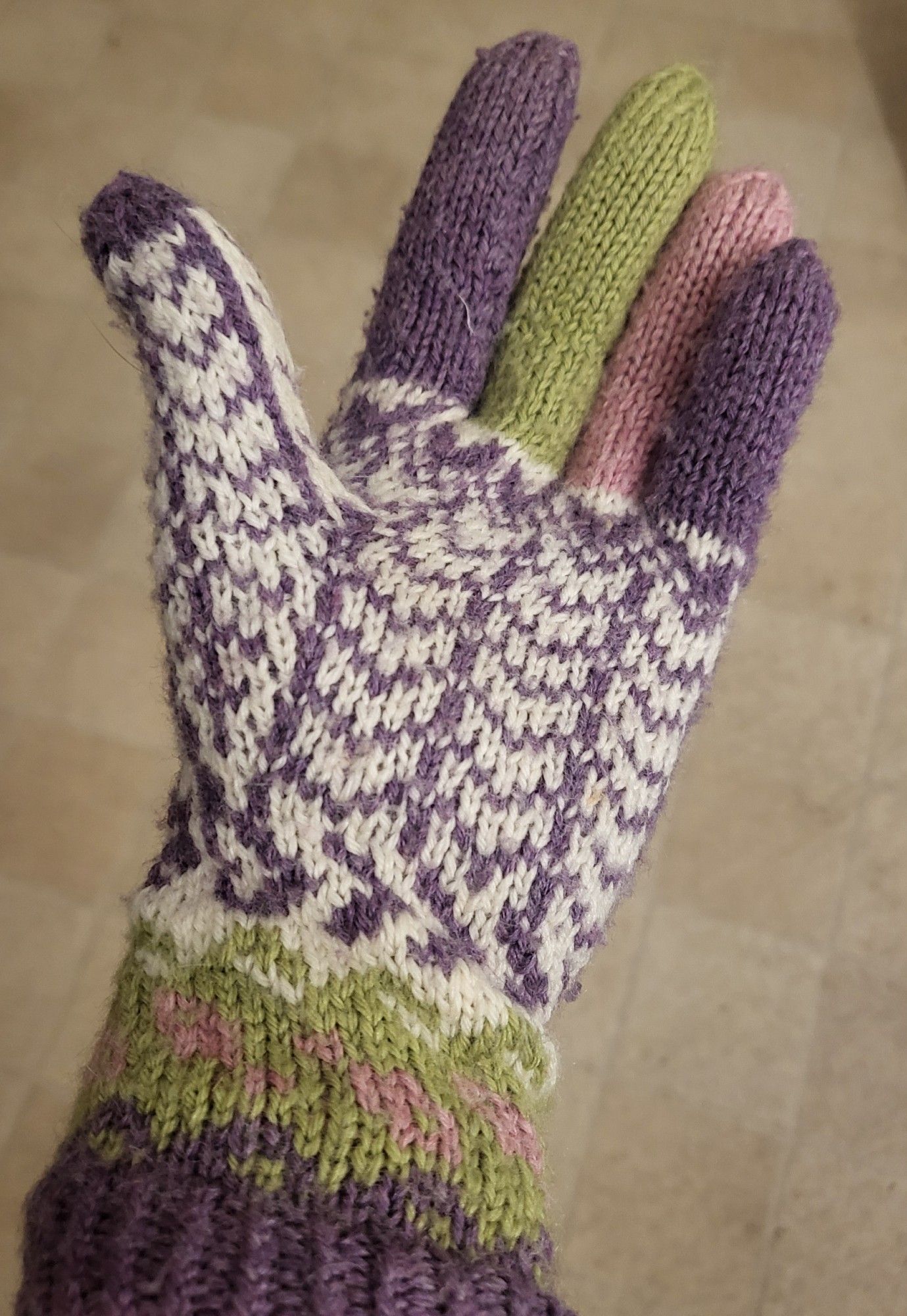 A handknitted glove with purple, green and pink fingers, and cuff, and the thumb/hand section in a purple ladder style pattern on a white background