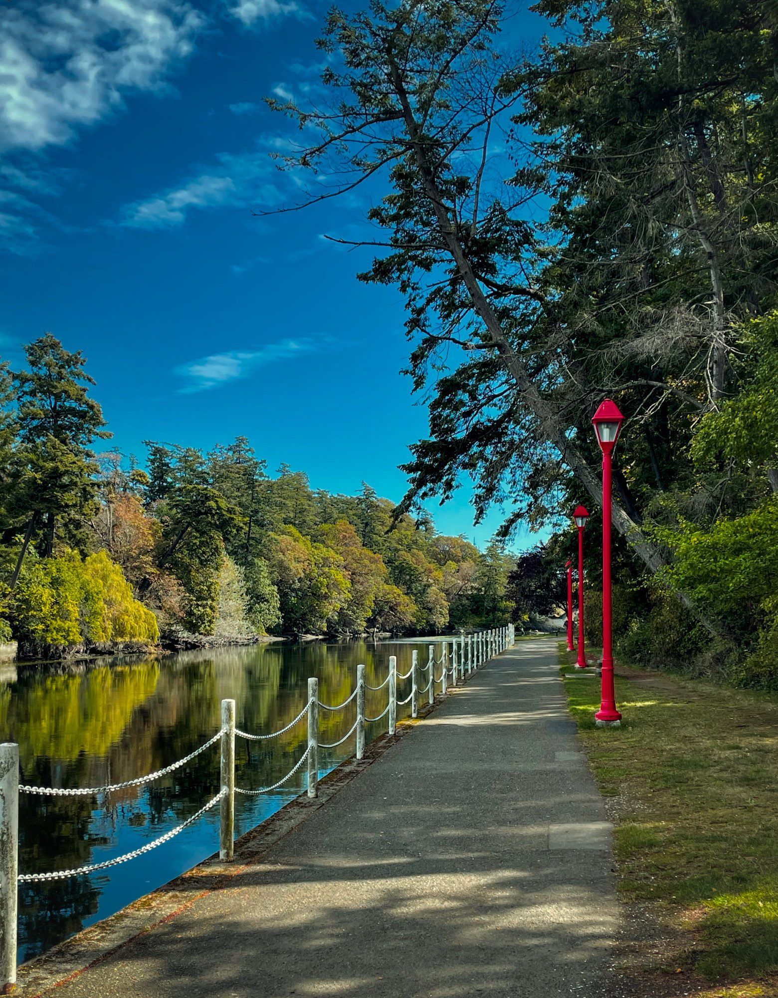 A paved pathway with chain fence bordering a waterway.  There are three tall red street lights lining the walkway.  Across the water are trees in various stages of change reflecting in the water.  The sky is bright blue with a few streaky clouds.