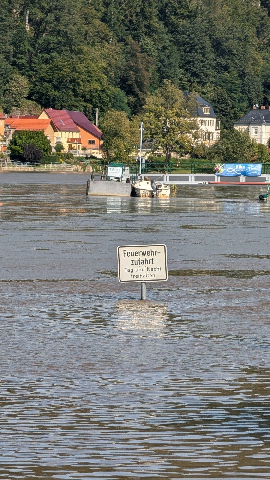 Auf dem Bild sieht man eine überschwemmte Straße. Ein Schild ragt aus dem Wasser heraus, auf dem 'Feuerwehrzufahrt, Tag und Nacht freihalten' steht. Im Hintergrund sind mehrere Häuser zu sehen, die am Ufer eines Flusses stehen. Der Fluss hat Hochwasser und hat Teile des Ufers und der Straße überflutet.