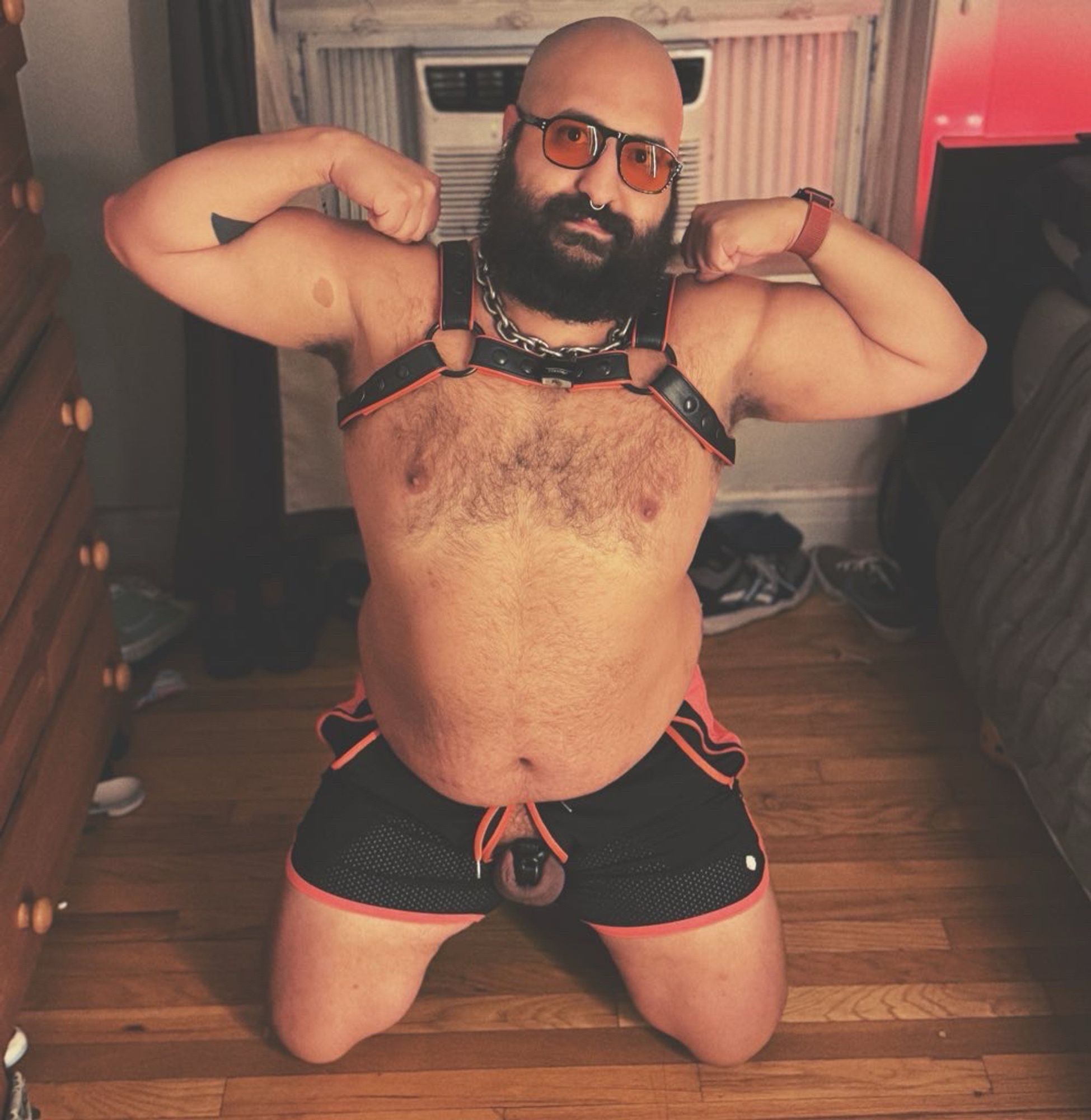 Photo of me kneeling wearing a harness and shorts. The shorts are open and my locked nub is exposed