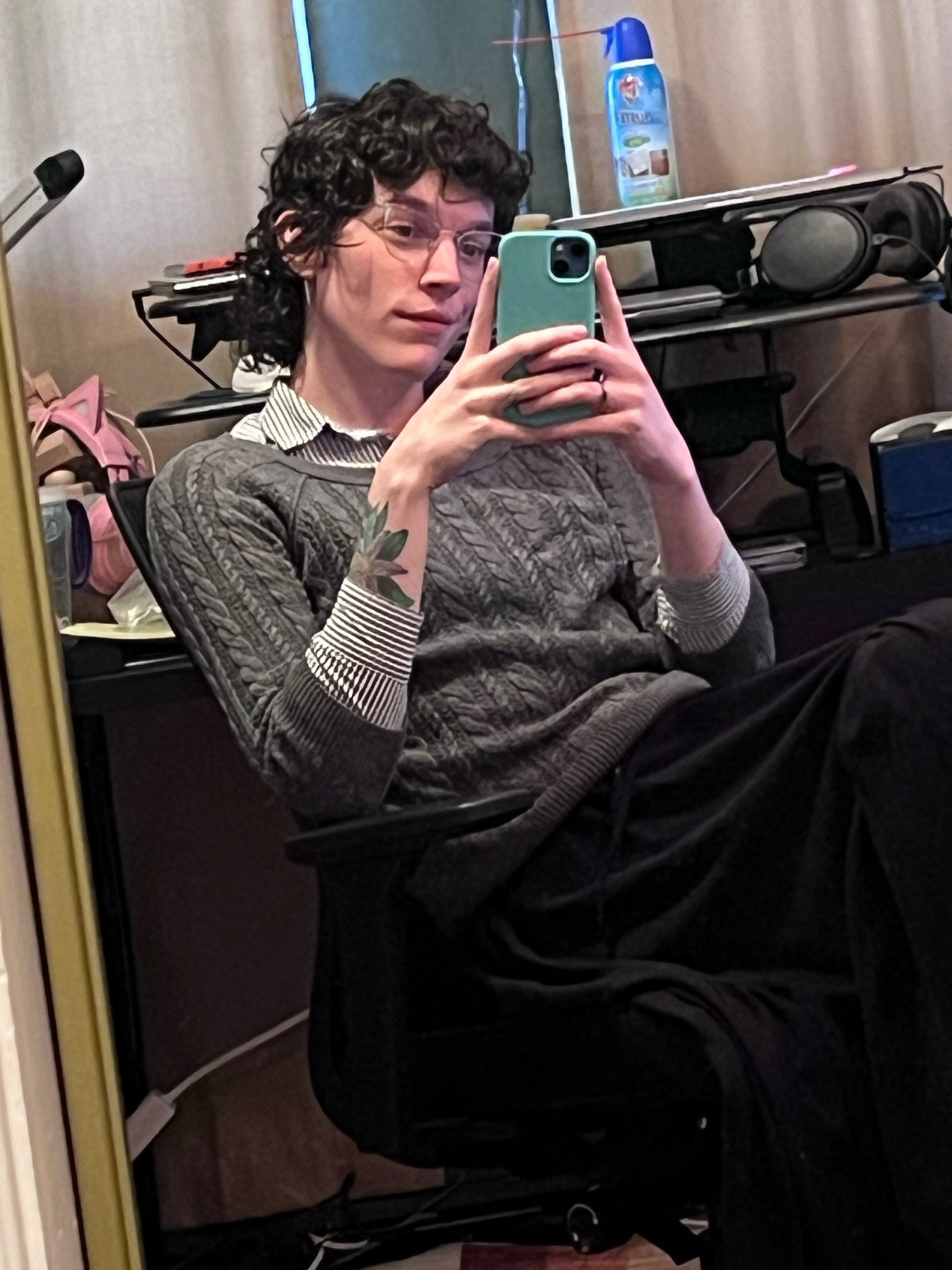 white enby reclining in their office chair, they have a grey sweater on over button down, and a long black skirt. They’re looking softly at their phone taking the picture