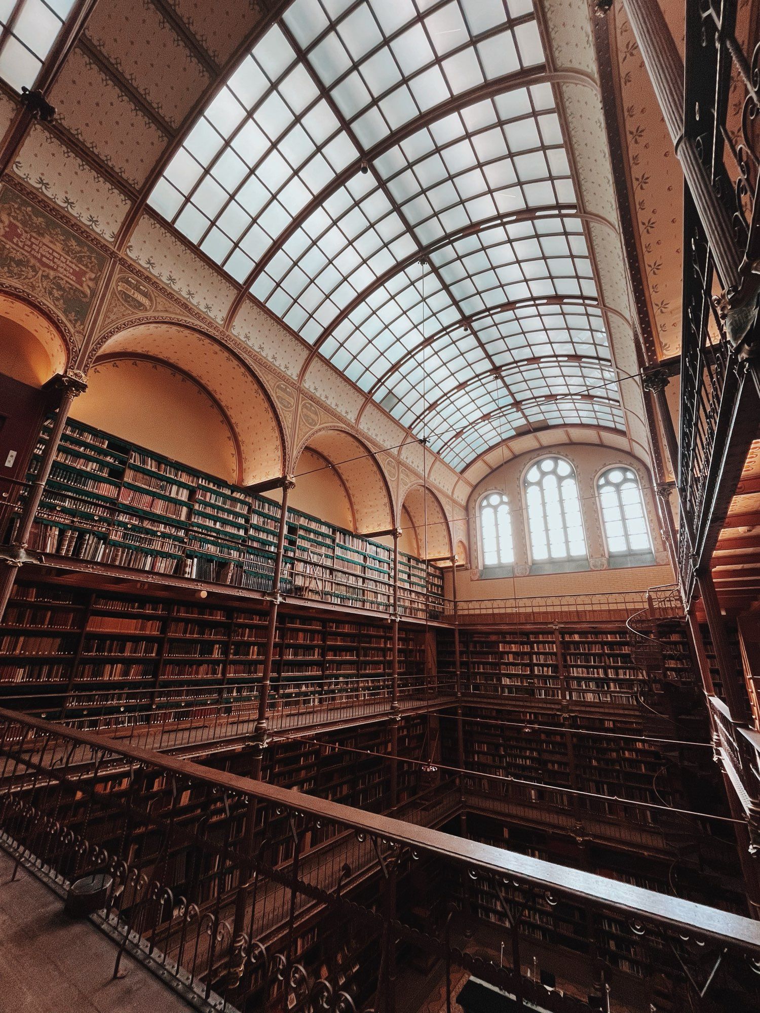 Een foto van de oude bibliotheek in het Rijksmuseum. Je staat op een hogere verdieping en kijkt neer op meerdere verdiepingen met rondom hoge boekenkasten gevuld met boeken. Het plafond is beschilderd. En valt licht door het in boog ronde glazen dak. En er zijn 3 statige hoge ramen, ook met bogen aan de bovenkant.