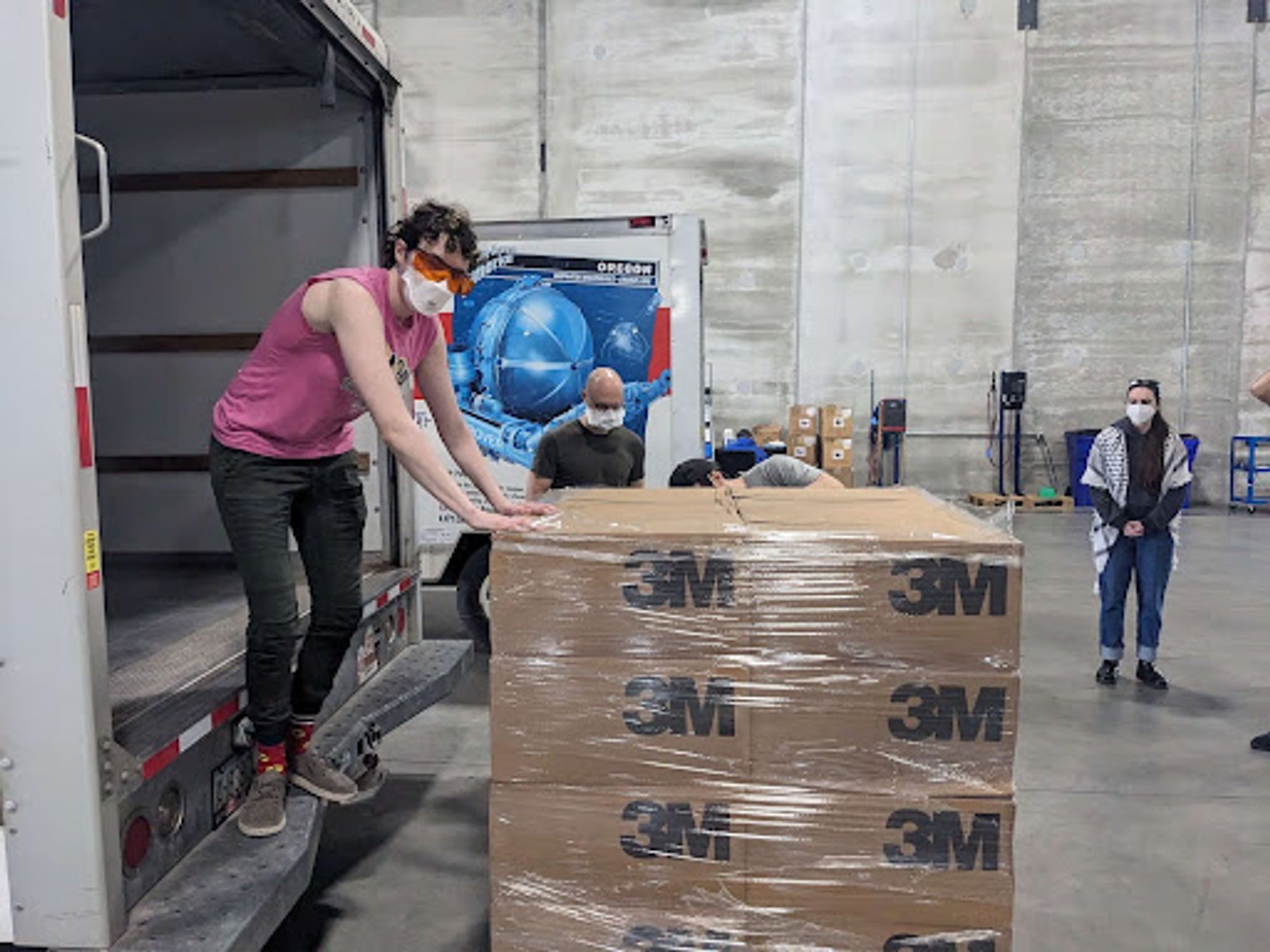 A person stands in a uhaul truck loading a pallet of masks into it. They are wearing an n95 mask and safety glasses.