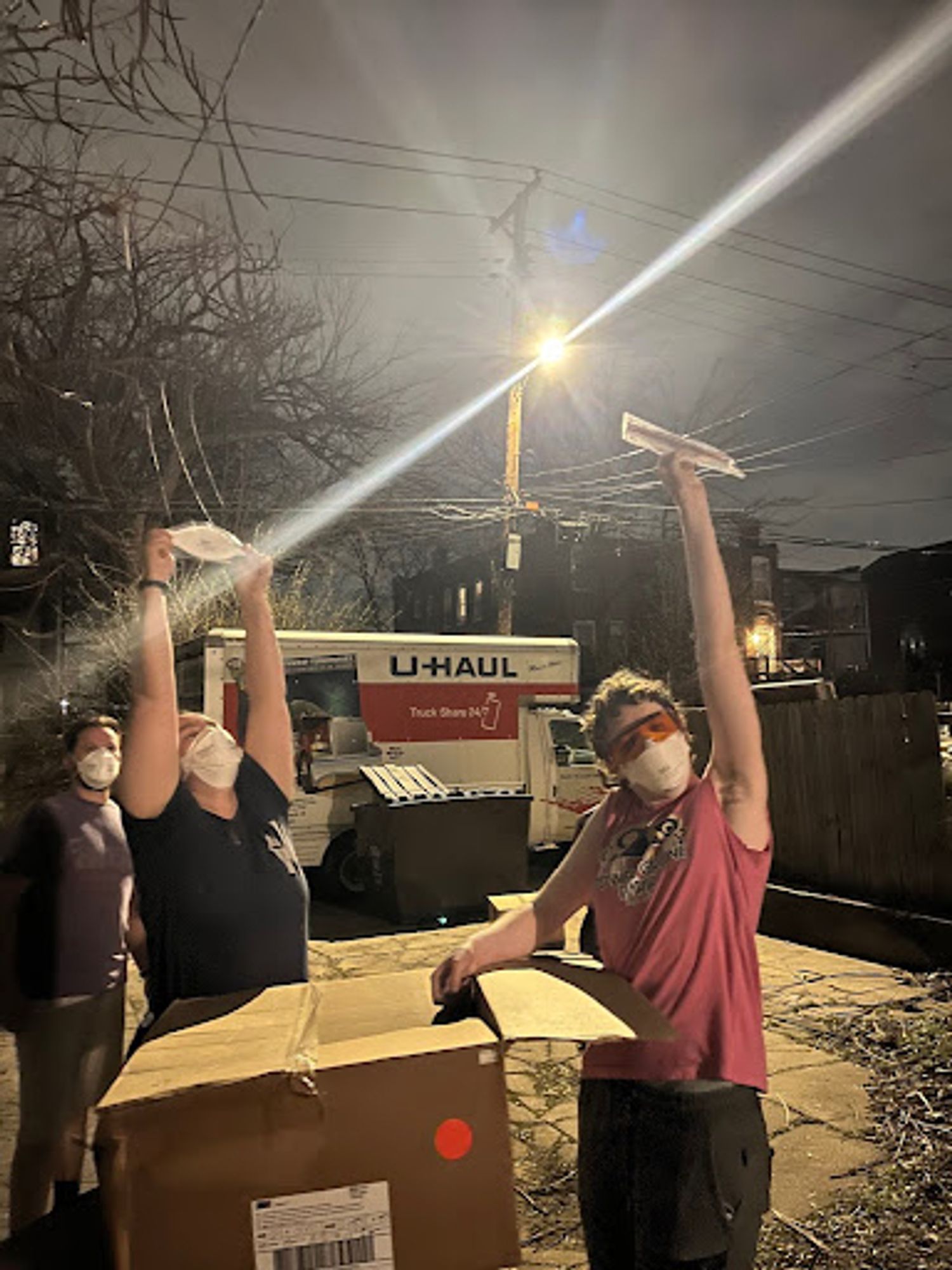 3 humans hold packaged n95s over their heads triumphantly. There is an empty uhaul in the image’s background and a full box of masks in the foreground.