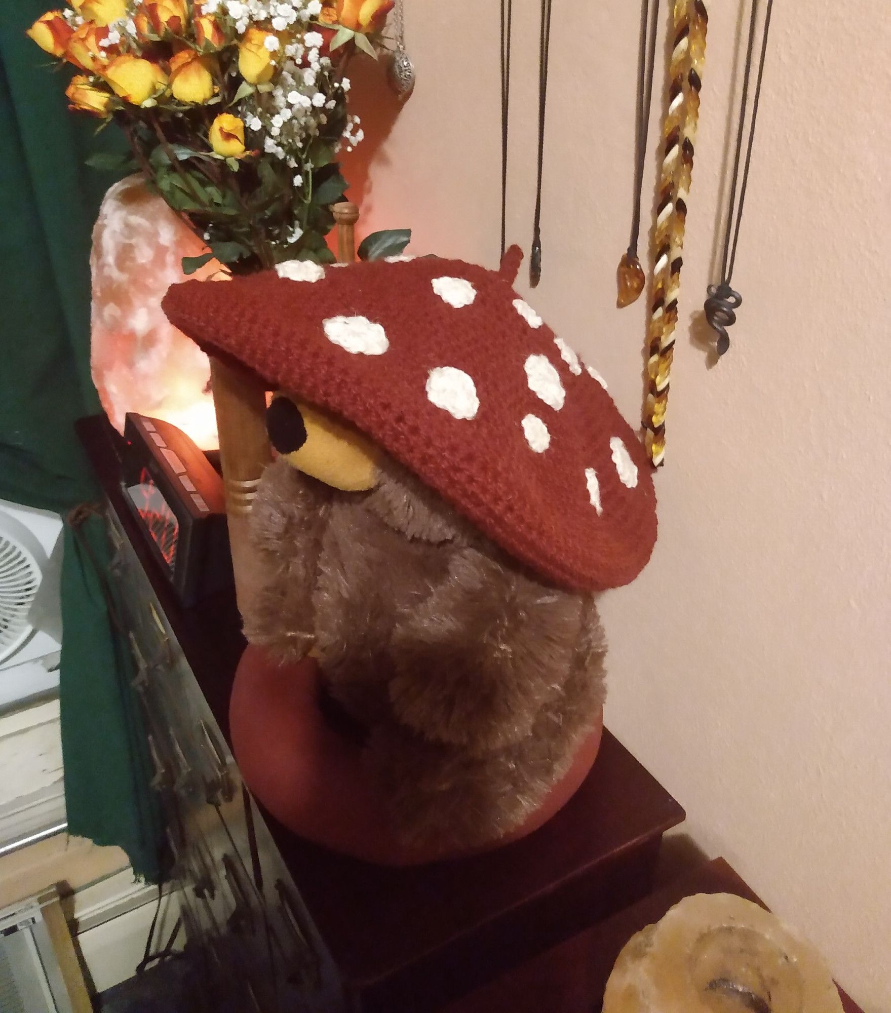 A stuffed bear named Humphrey sitting on a pillow ring meant for holding  a singing bowl. He is wearing a toadstool style crocheted beret that is red and covered in white spots, it is oversized and you can not see his eyes. in the back ground there are yellow roses tipped in orange, a salt lamp, alarm clock, and some necklaces hanging on the wall