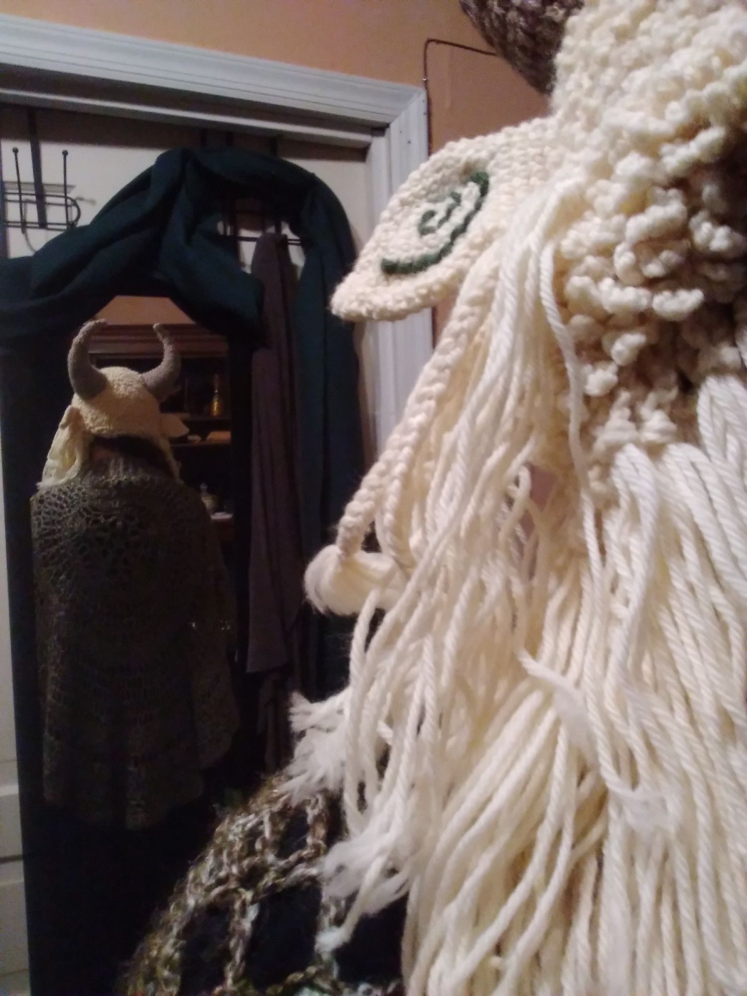 a person wearing a white goat hat that has ears with green spirals on them, brown horns, and lot of shaggy goat hair with some braids in it. you don't see their face but you do see the back of the hat in a mirror (also wearing a green crochet cape/poncho thing)