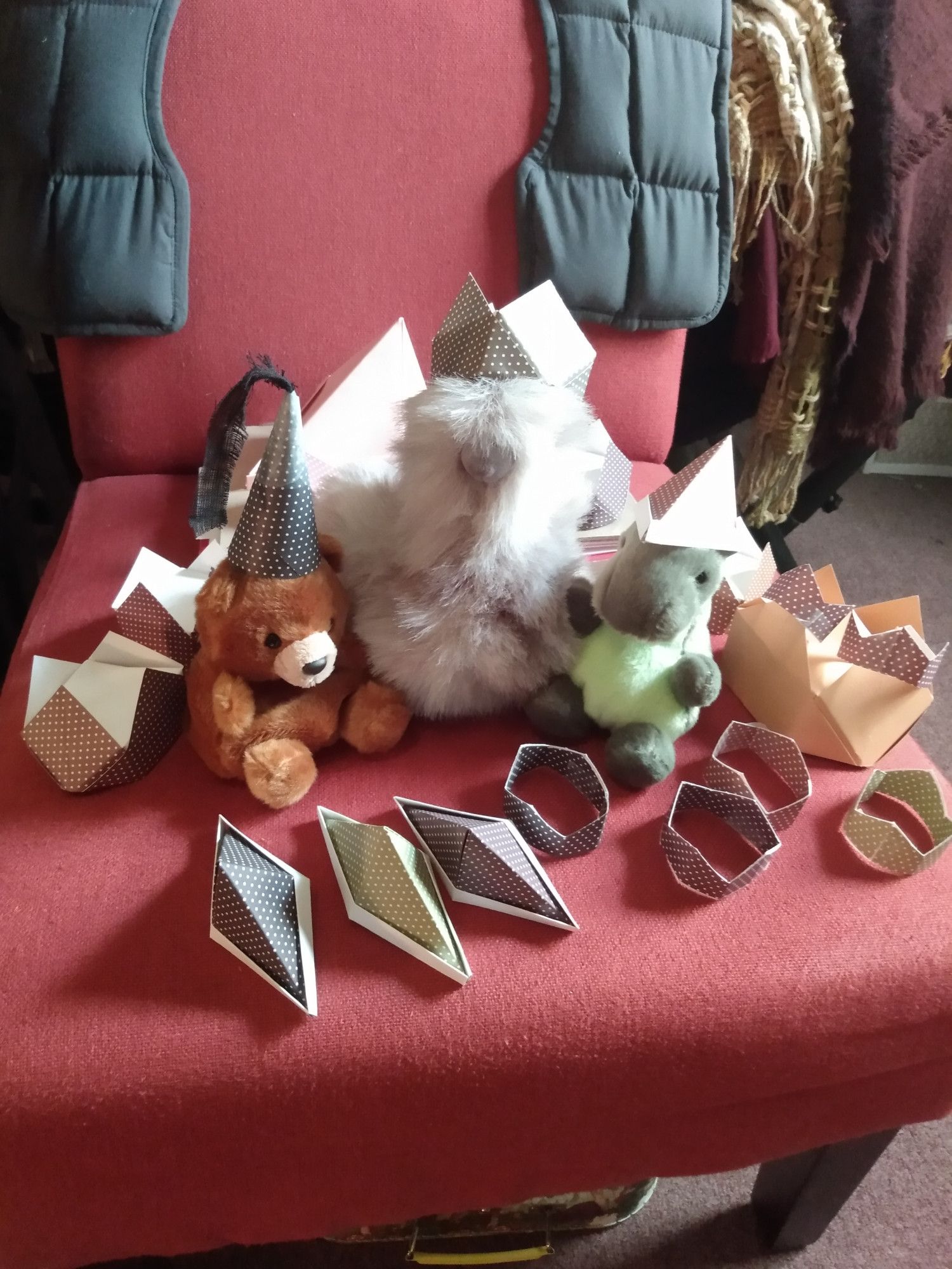 three stuffed animal sitting on a chair surrounded by various folded paper hats. They are also wearing paper hats. to the left is Arthur a bear, he is wearing a conical "princess hat" that is to say a single hennin, in the center is a grey bird they are wearing a square crownish origami hat, to the left is Martha a T-rex style dinosaur wearing ye old newspaper hat/boat everyone used to make.