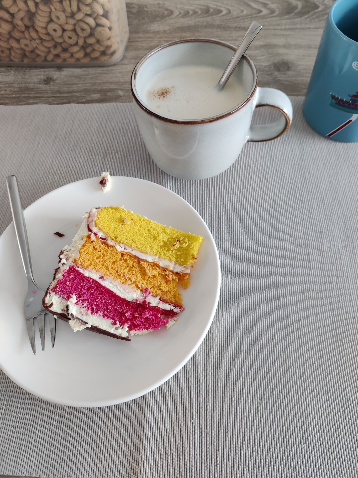 Een stuk dripcake van Hema op een bordje (roze oranje en geel) en een mok latte macchiato op een grijs placemat (met nog een stukje blauwe Efteling mok te zien ernaast rechts)