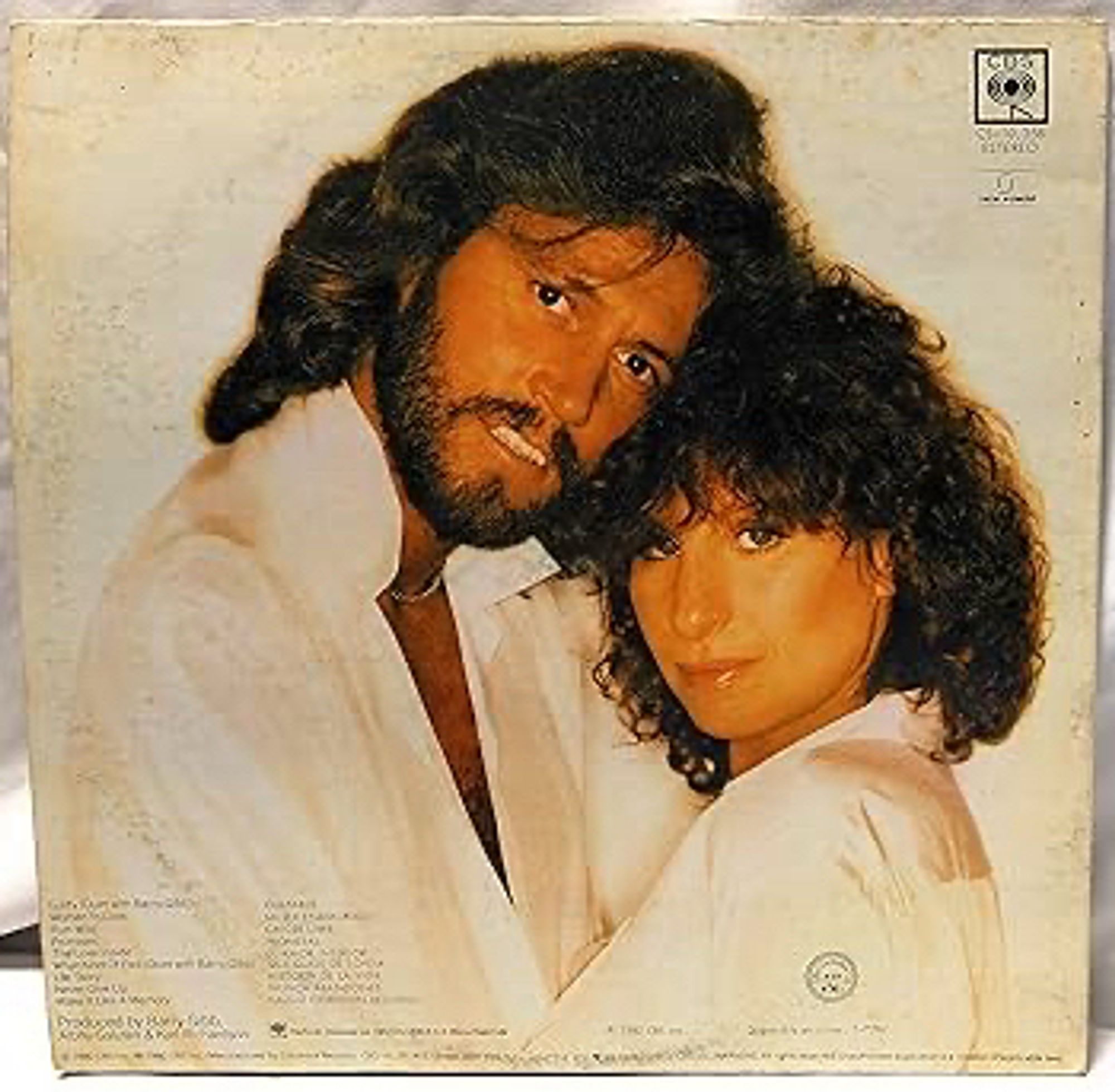 The back cover of the Barbra Streisand album guilty from 1980. Barbra is shown with Barry Gibb from the Bee Gees. Barbra and Barry are hugging - wearing white on a white background - staring wide-eyed down the camera lens, pupils huge, eyeballing the viewer. Barbra smoulders in a sleepy way. Barry has a clenched teeth grin.