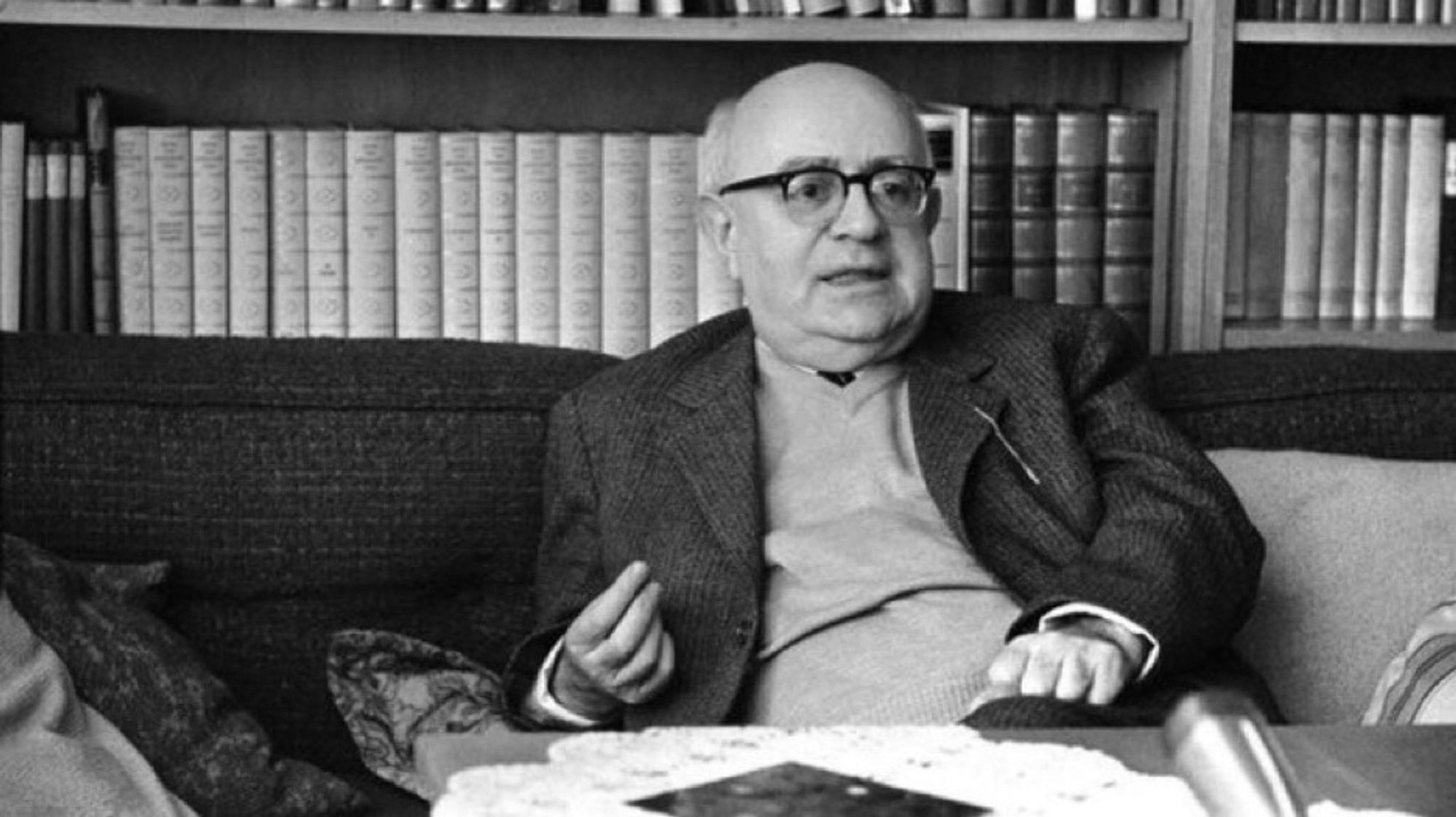 Adorno sitzt mit Pulli und Jacket vor seine Bücherwand. Er schaut einen Betrachter an und redet wohl gerade.