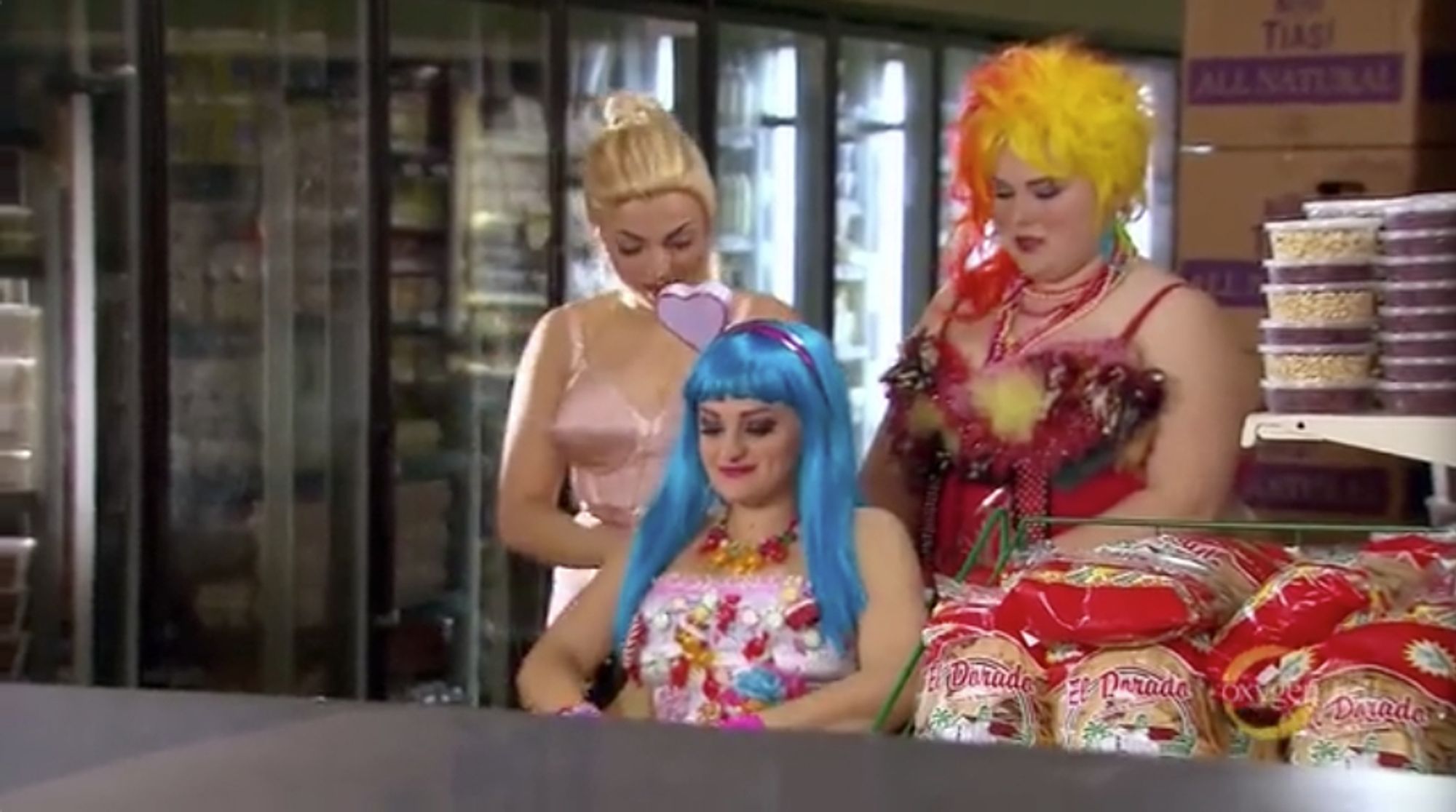 Aylin, Ali, and Lily Mae from the Glee Project are in a supermarket at night. Aylin is dressed like Madonna (blonde, cone bra), Ali is dressed like Katy Perry (blue wig and candy dress), and Lily Mae is dressed like Cyndi Lauper (80s her and feathery dress).