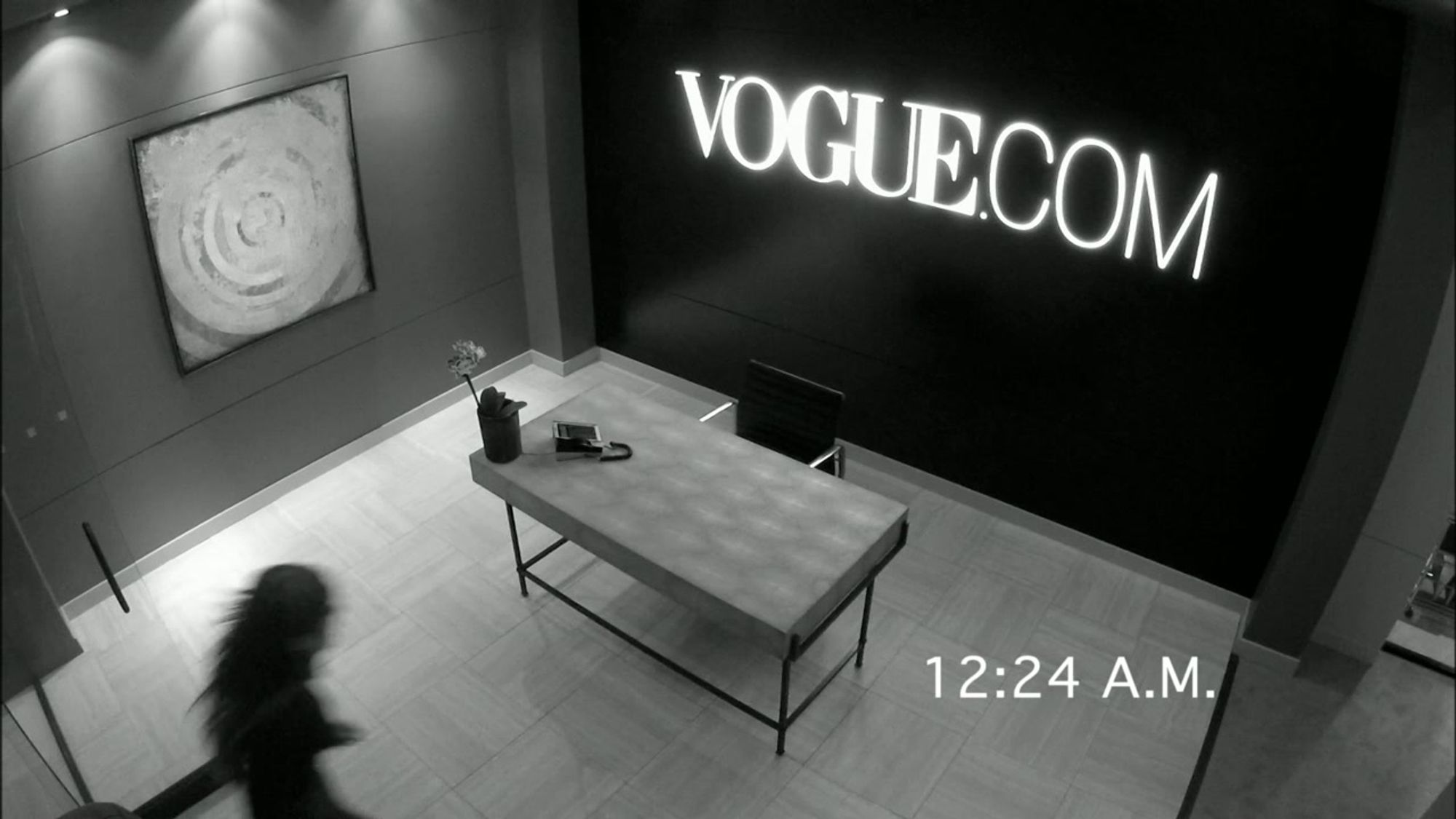 Black and white security camera footage of the Vogue.Com offices. It's 12:24 AM, and a blurry Rachel Berry is scurrying into the offices.