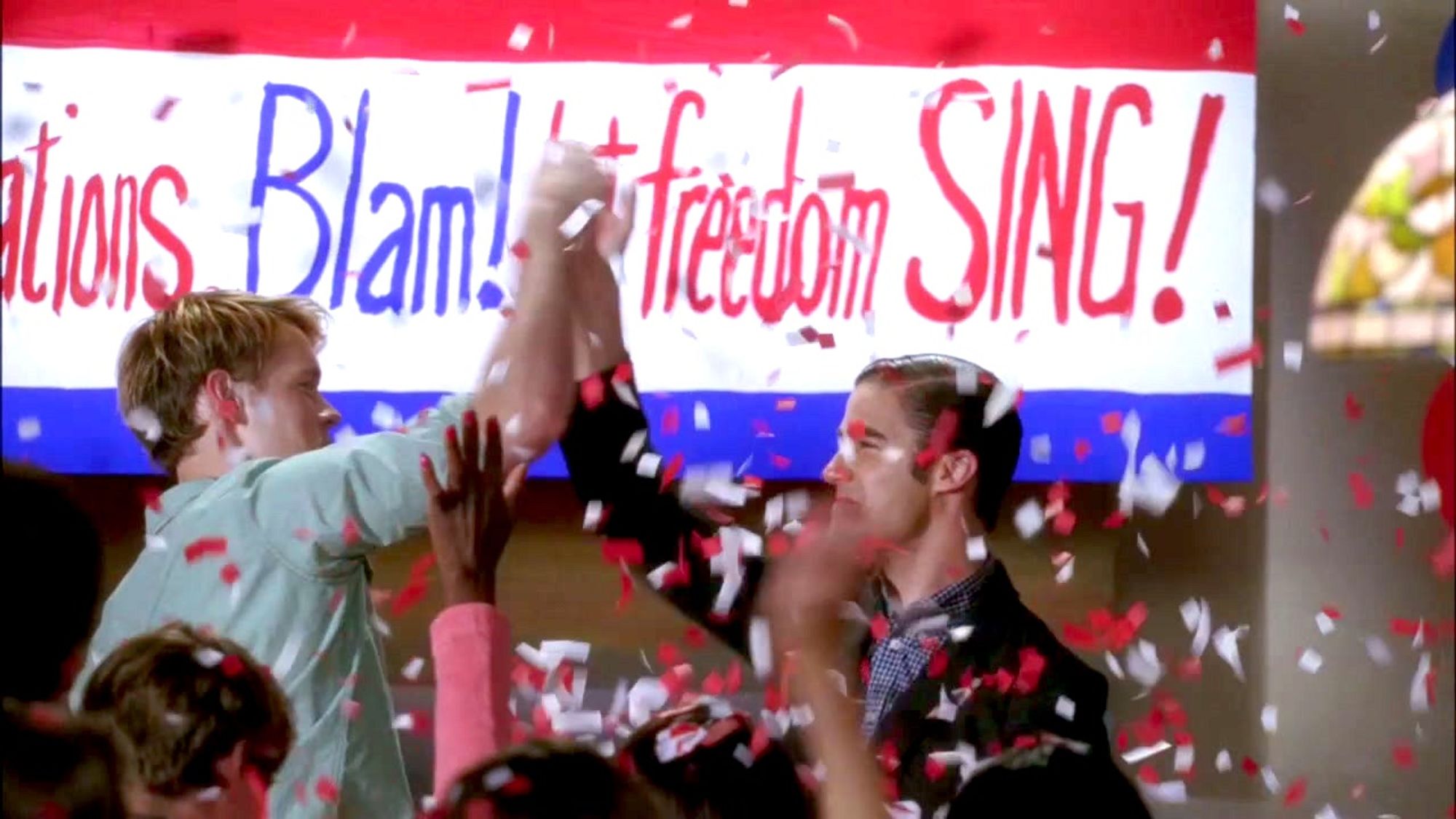 Sam and Blaine from Glee hi-five excitedly at a party for winning student elections. Confetti rains down, and behind them is a banner that reads "Congratulations Blam! Let freedom SING!"