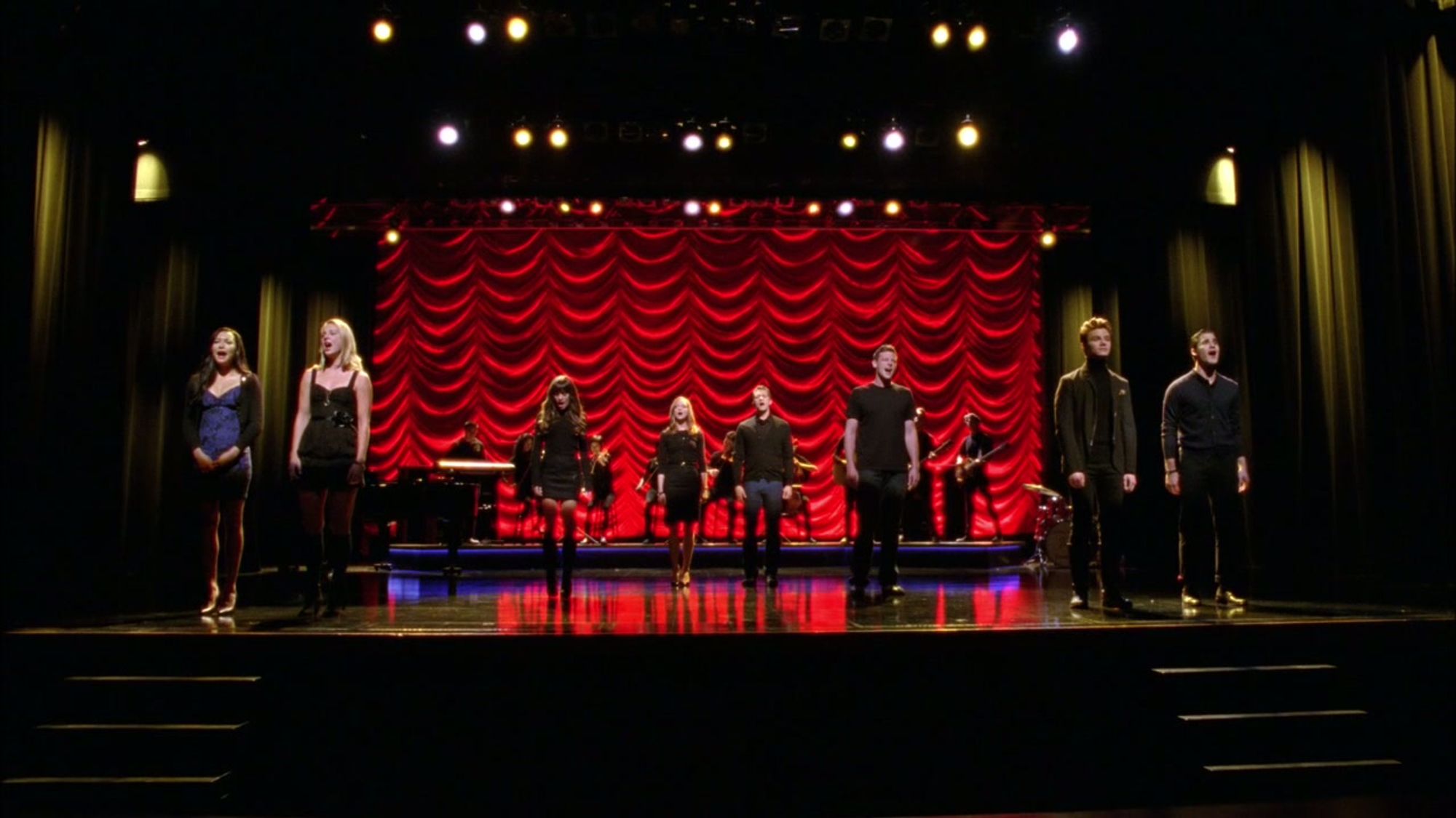 Four couples from Glee (Santana and Brittany, Rachel and Finn, Emma and Will, Kurt and Blaine) stand next to each other on stage in front of a red curtain, staring out to the audience and making it clear from body language that there is a distance between the members of each couple.