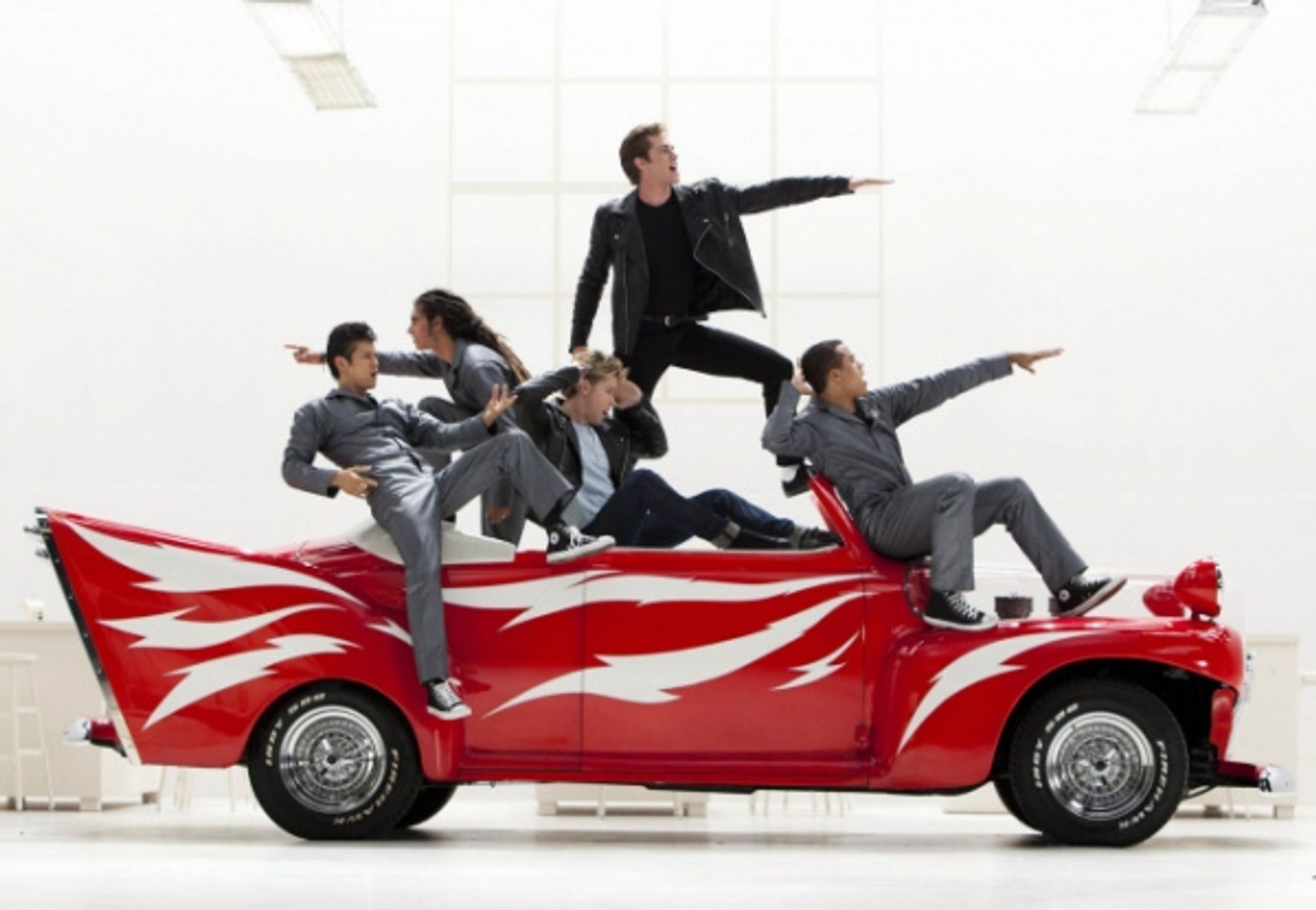 Mike, Joe, Sam, Ryder, and Jake from Glee pose atop a red hot rod with white streaks, wearing silver jumpsuits similar to the original "Greased Lightning" musical number.