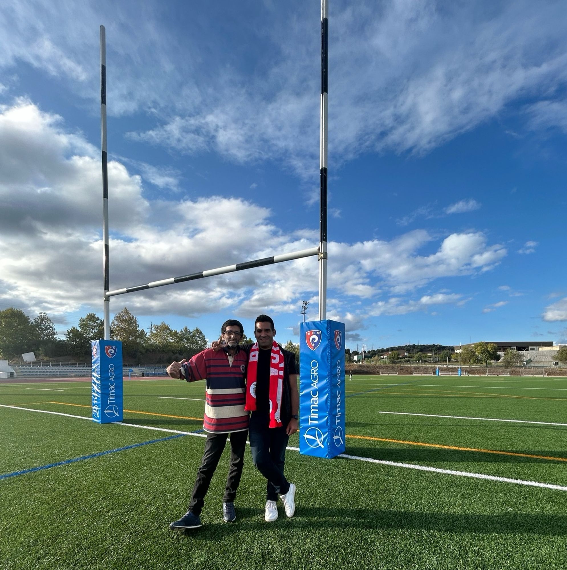 Two people standing on a rugby field near goalposts, smiling and posing against a backdrop of a cloudy sky. One person wears a striped shirt, the other a scarf.