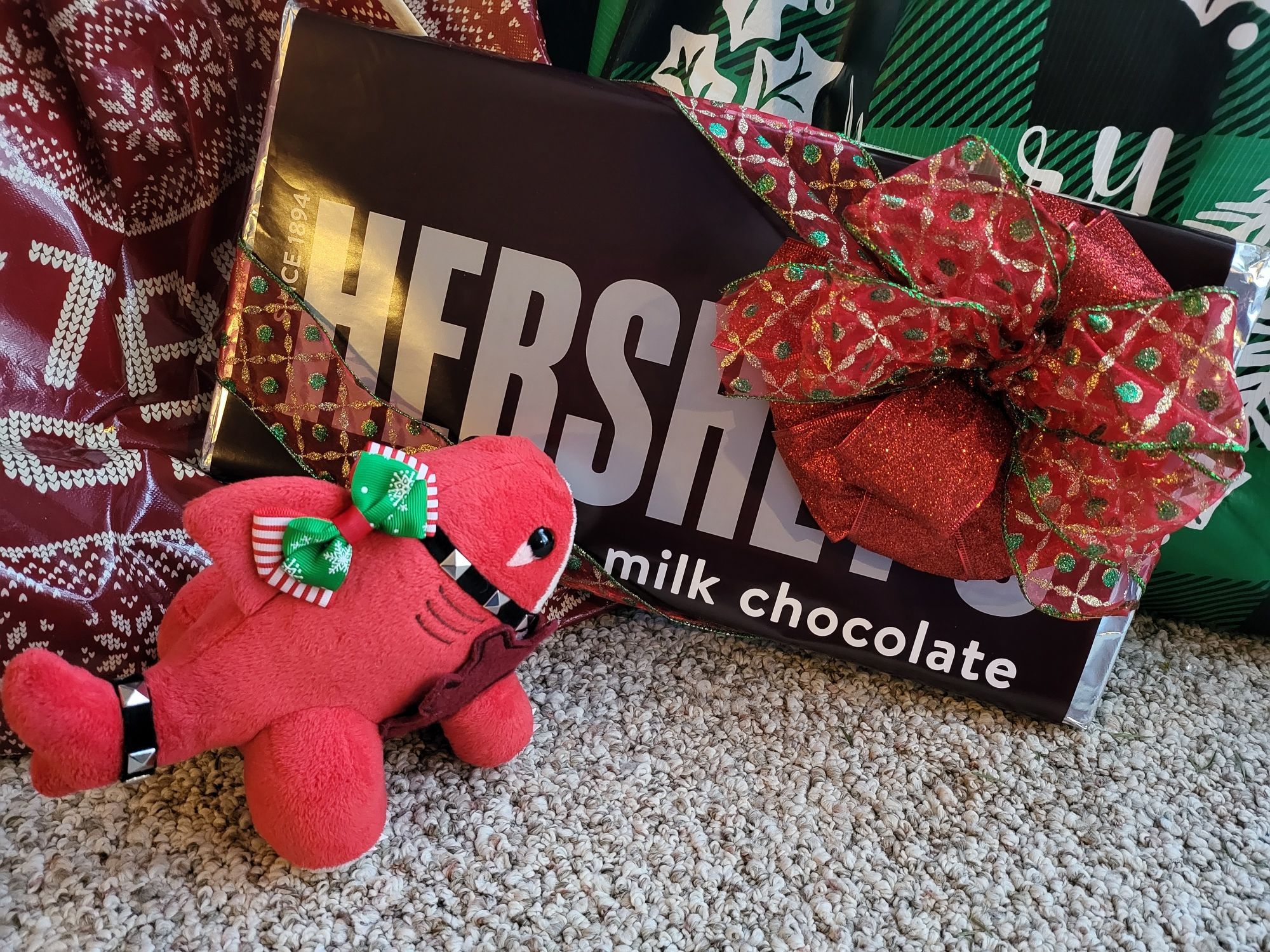 a little red landshark plush wearing a festive bow on his fin stands in front of a gift-wrapped chocolate bar that is four times as big as him with a very determined expression.