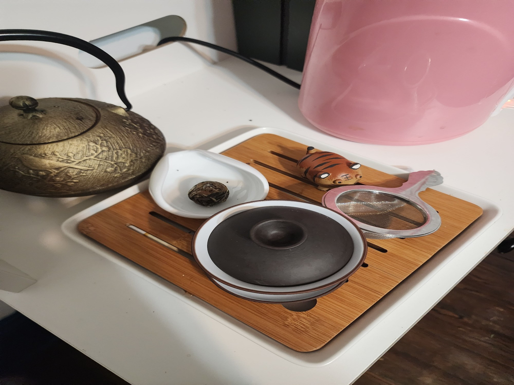 Gong fu Tea set with a dark colored gaiwan, a white tray containing a ball of compressed white tea leaves, a strainer, and a fat tiger tea pet.