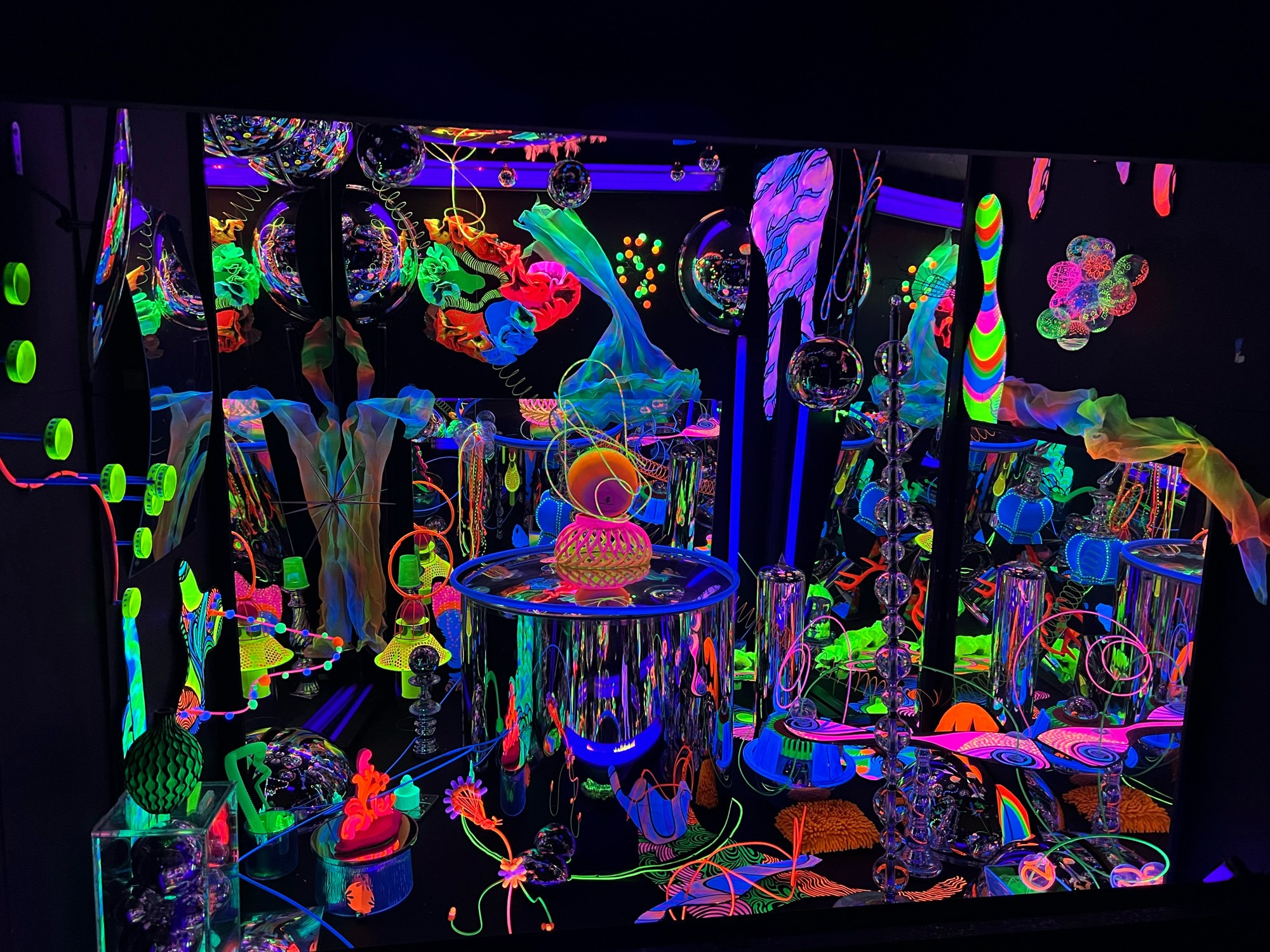 An art piece consisting of an assemblage of random shapes and objects painted with bright glowing paint (orange, purple, lime green, pink, red). The effect is blobs of glowing color in a black background